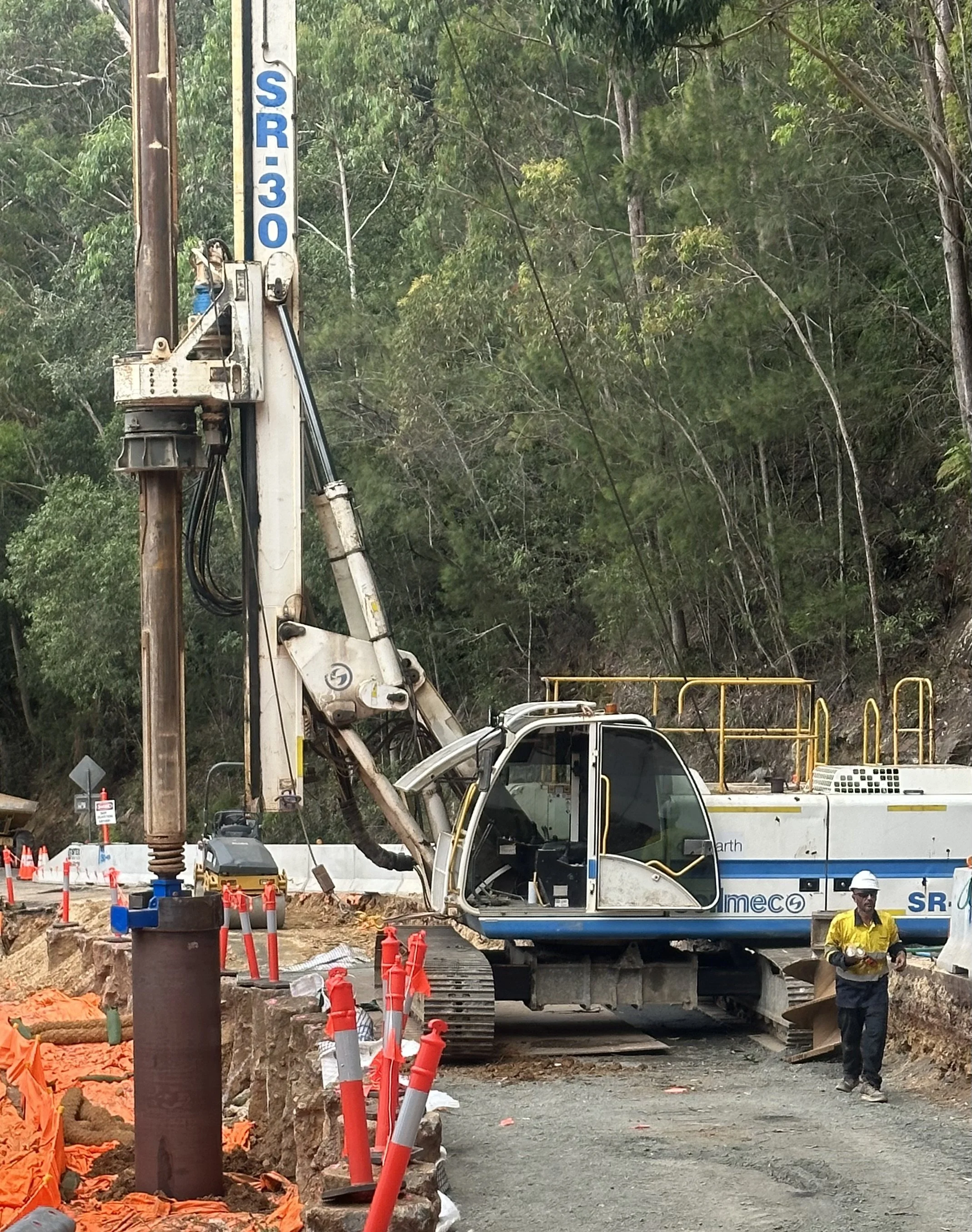 A construction site with a large drilling machine next to a dirt road, several orange and gray traffic cones, and a construction worker wearing a yellow safety vest and white hard hat.