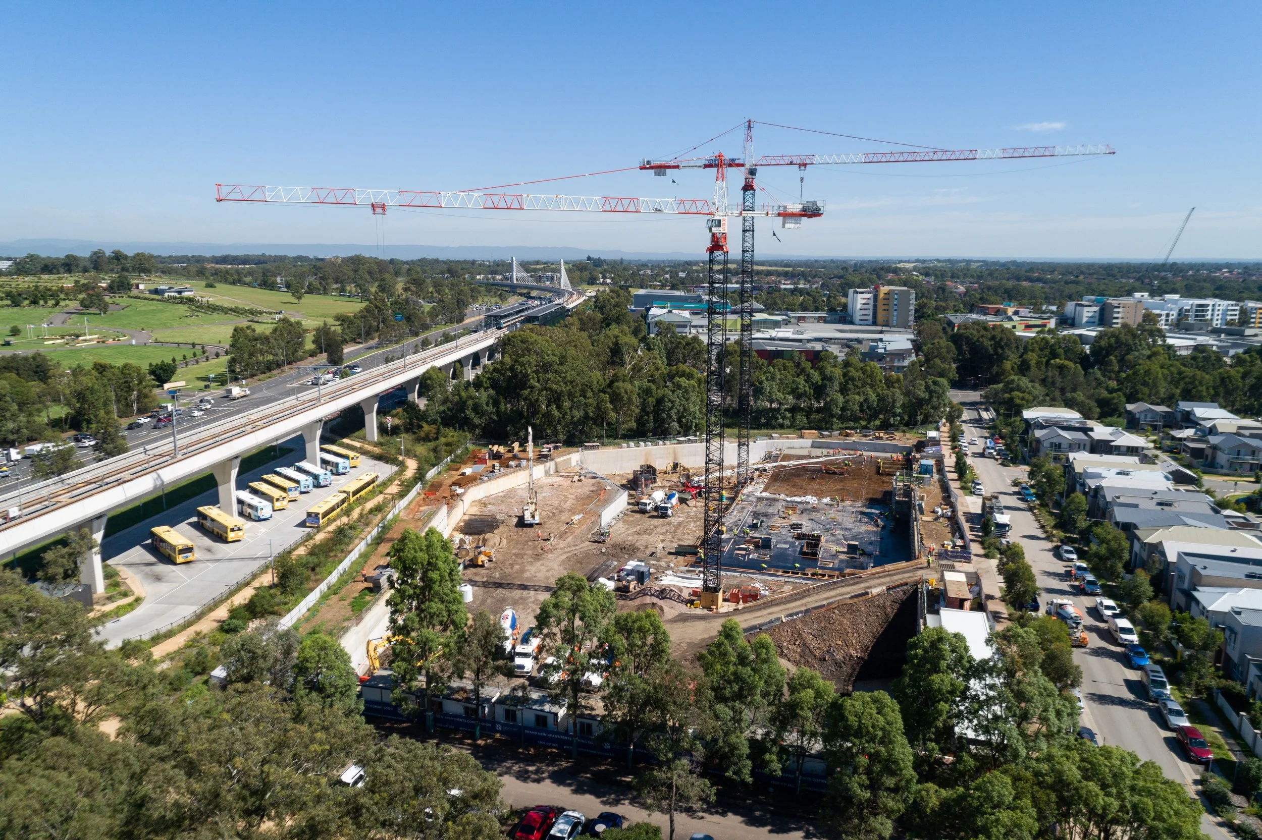 A construction site with a large crane, several smaller cranes, and heavy machinery, adjacent to a road with parked buses, in a suburban area with trees and buildings in the background.