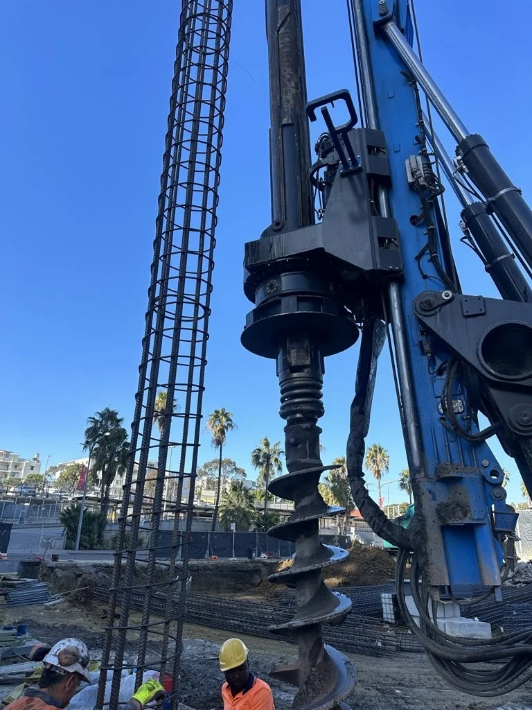 Construction site with a large blue drill and workers wearing hard hats, palm trees in the background.