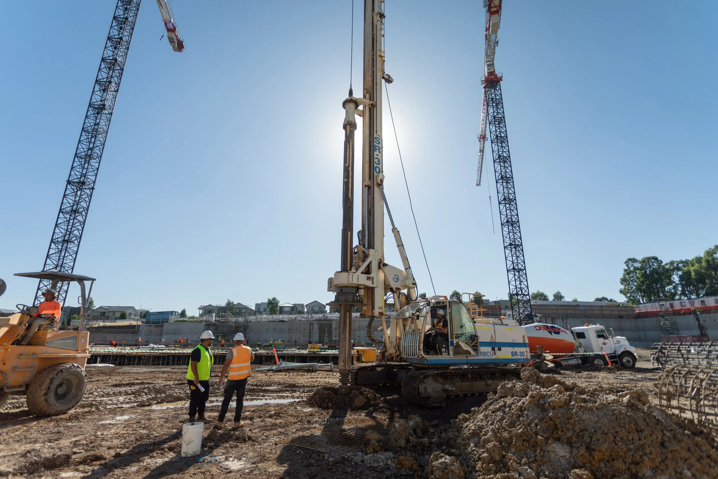 Construction site with a large crawler crane drilling into the ground, workers in safety vests and helmets, and two tall tower cranes against a clear blue sky.