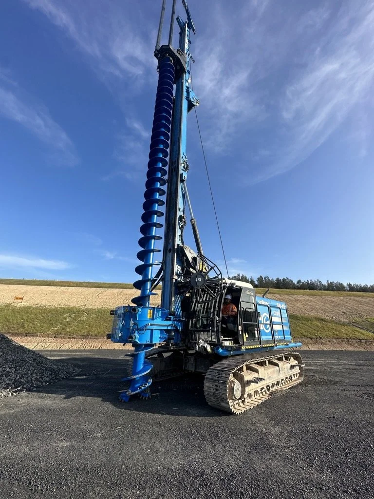A large blue drilling machine with a tall auger drill bit, tracked vehicle, and operator in the cab, on a construction site with dirt and grassy hills in the background under a partly cloudy sky.