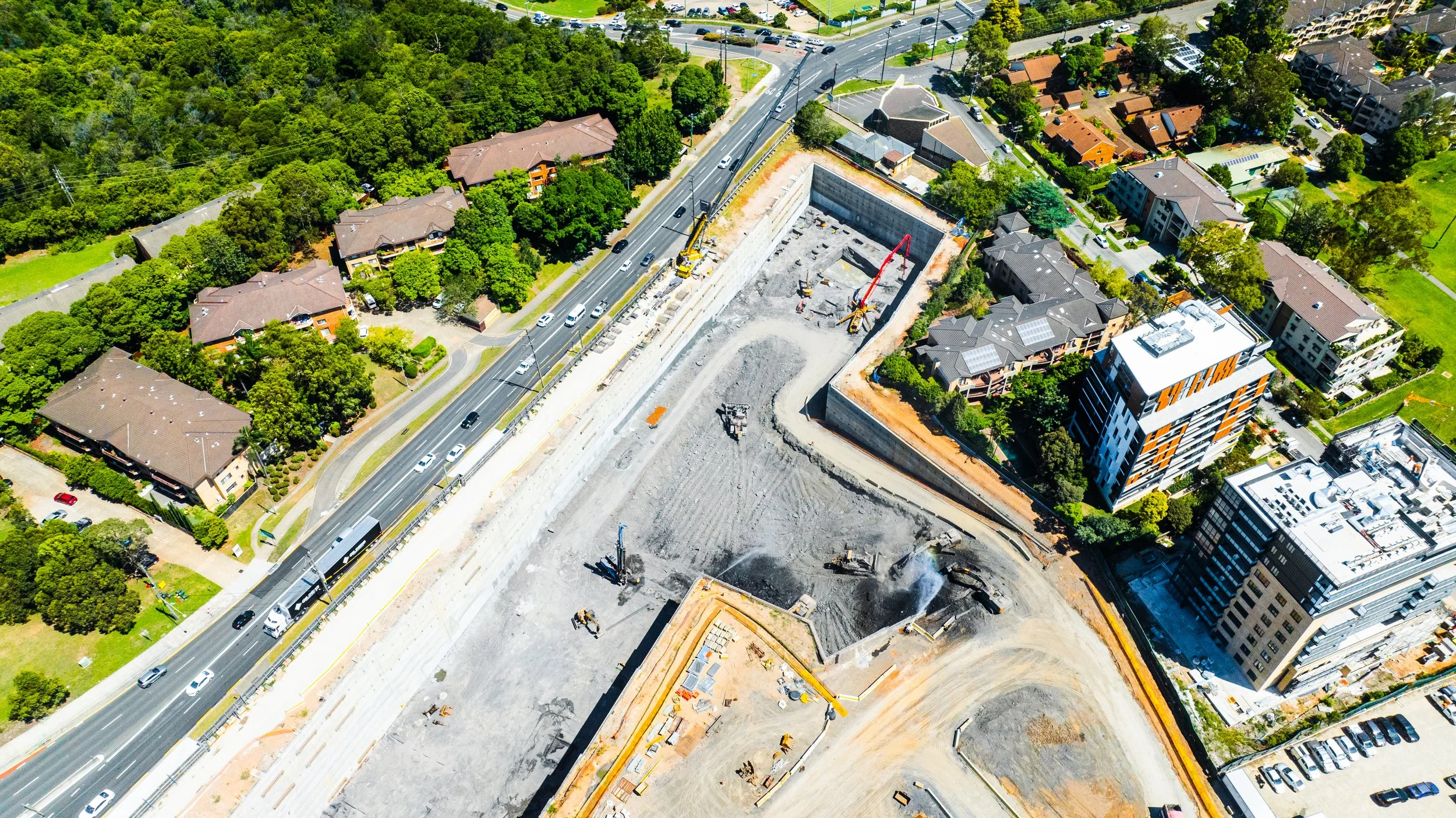 An aerial view of a construction site in an urban area with high-rise buildings and residential neighborhoods, showing excavation and building activities with construction equipment and vehicles.