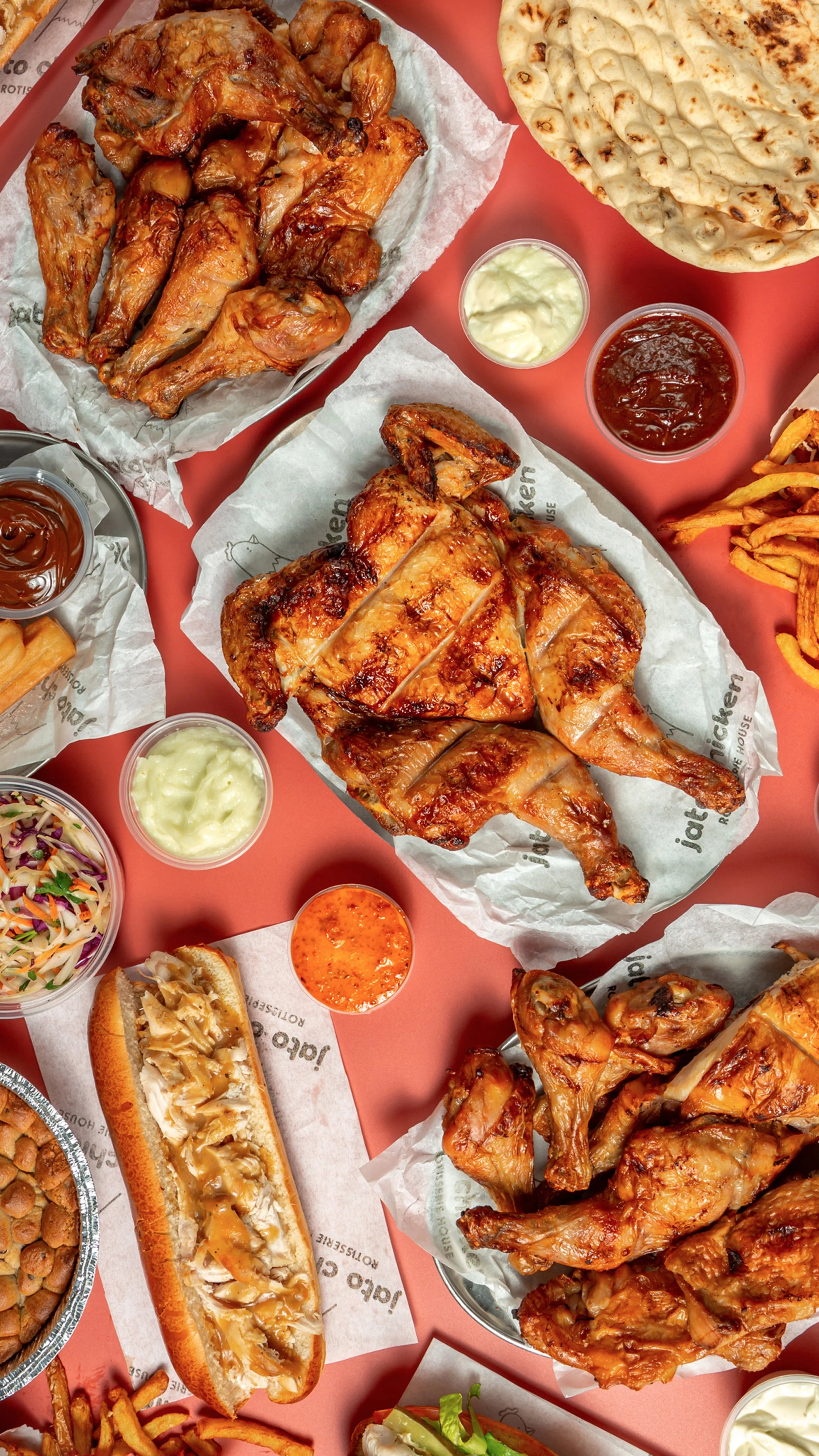 A variety of American fast food items including fried chicken, flatbread, fries, coleslaw, barbecue sauces, and a bacon cheeseburger, arranged on a pink table.