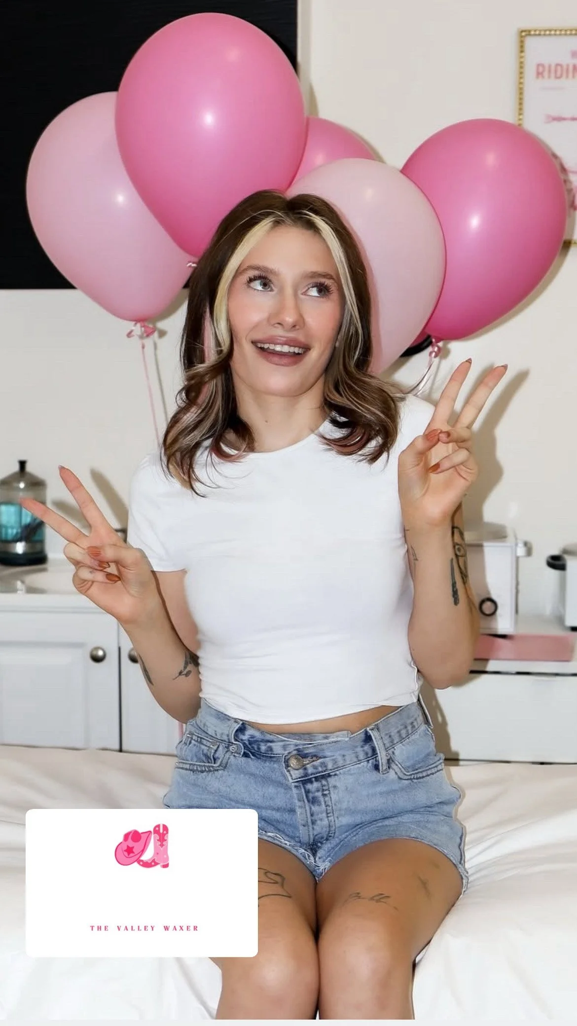 A young woman sitting on a bed, smiling, making peace signs with both hands, surrounded by pink balloons in the background, wearing a white crop top and denim shorts.