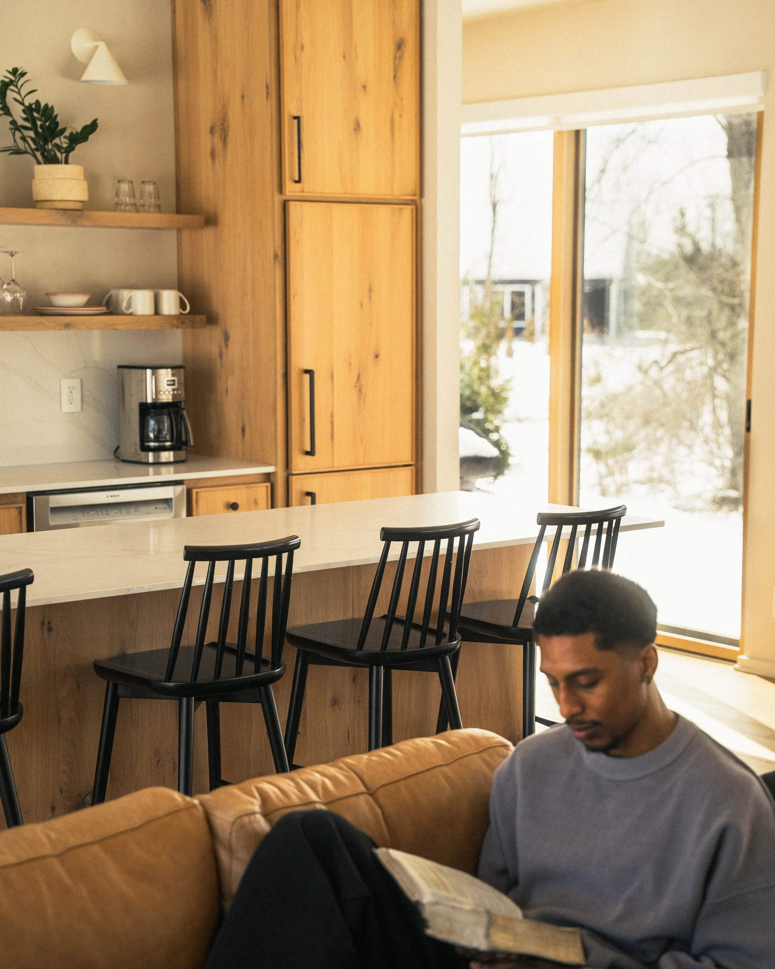A young man with dark hair and glasses sitting on a tan leather couch, reading a book, with a coffee table in front of him and black chairs in the background. There are large windows letting in natural light, and a wooden cabinet and shelves with dis