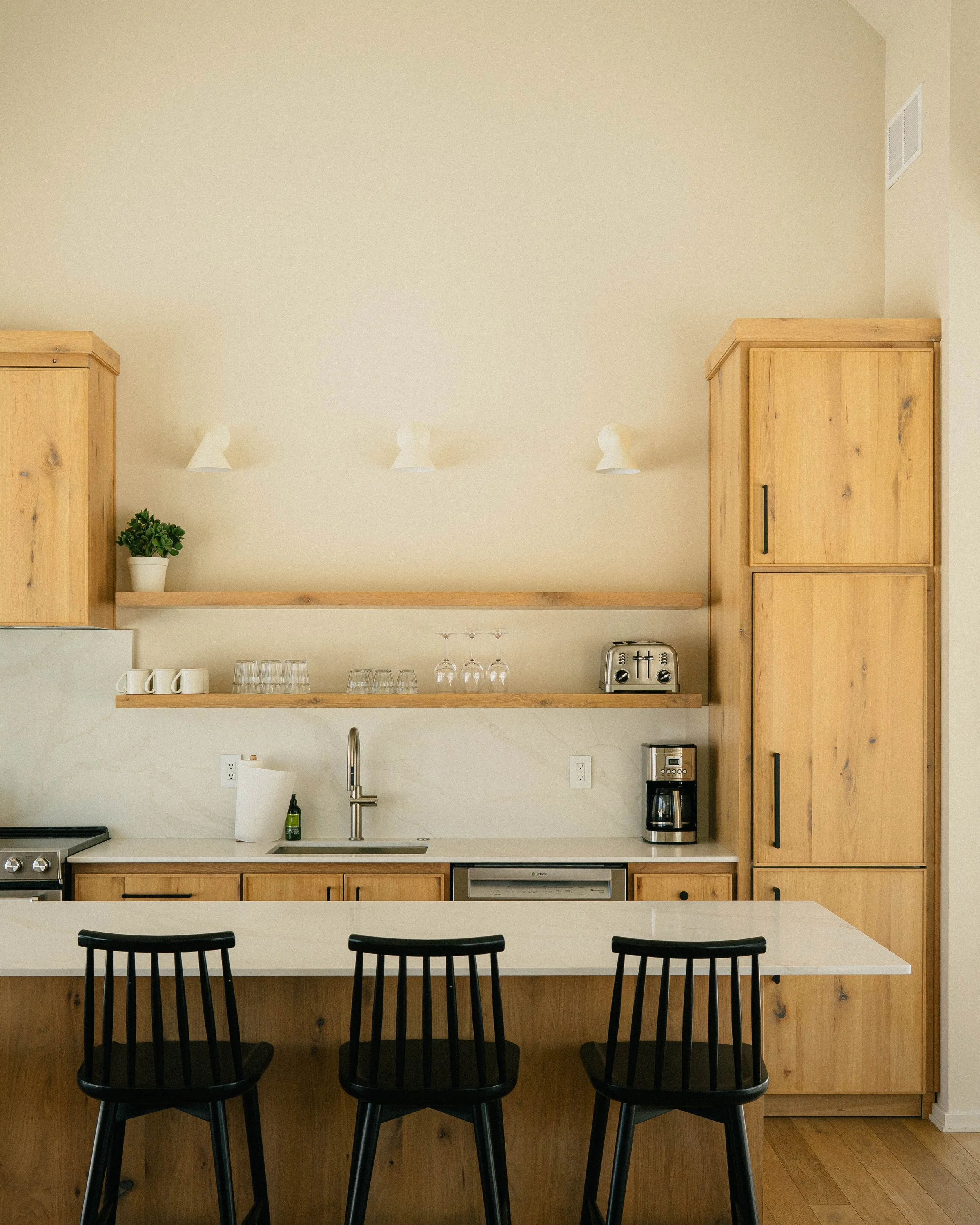 Kitchen with a white countertop, black chairs, wooden cabinets, and small appliances including a coffee maker and toaster. There are three barstools with black backrests in front of the counter.