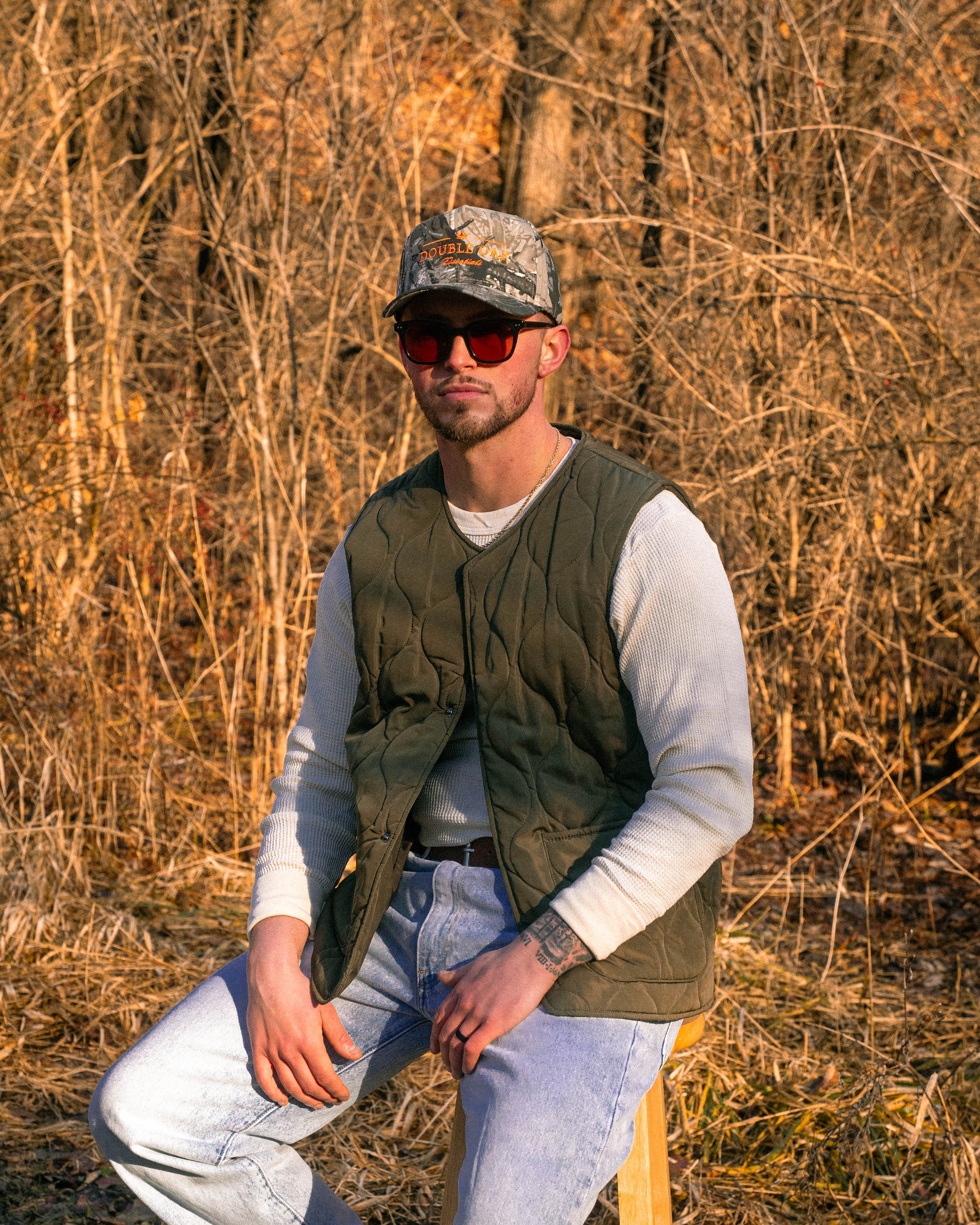 A man sitting outdoors in a wooded area with brown autumn leaves in the background, wearing a camouflage cap with orange text, dark sunglasses, a white long-sleeve shirt, and a green vest, with tattooed hands resting on his knee.