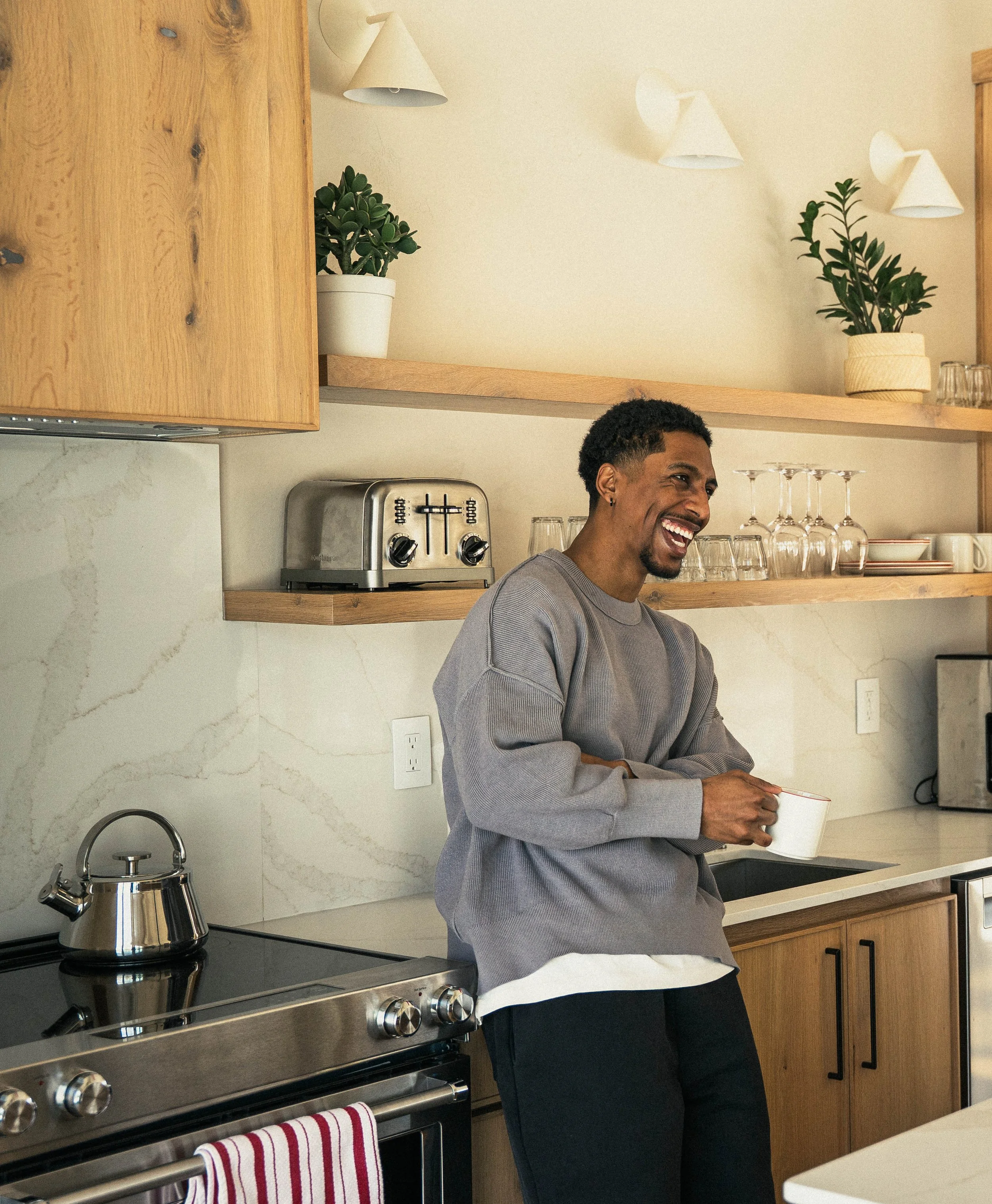 A man smiling and holding a mug in a modern kitchen with wooden cabinets, a stainless steel stove, plants on open shelves, and white cone-shaped wall sconces.