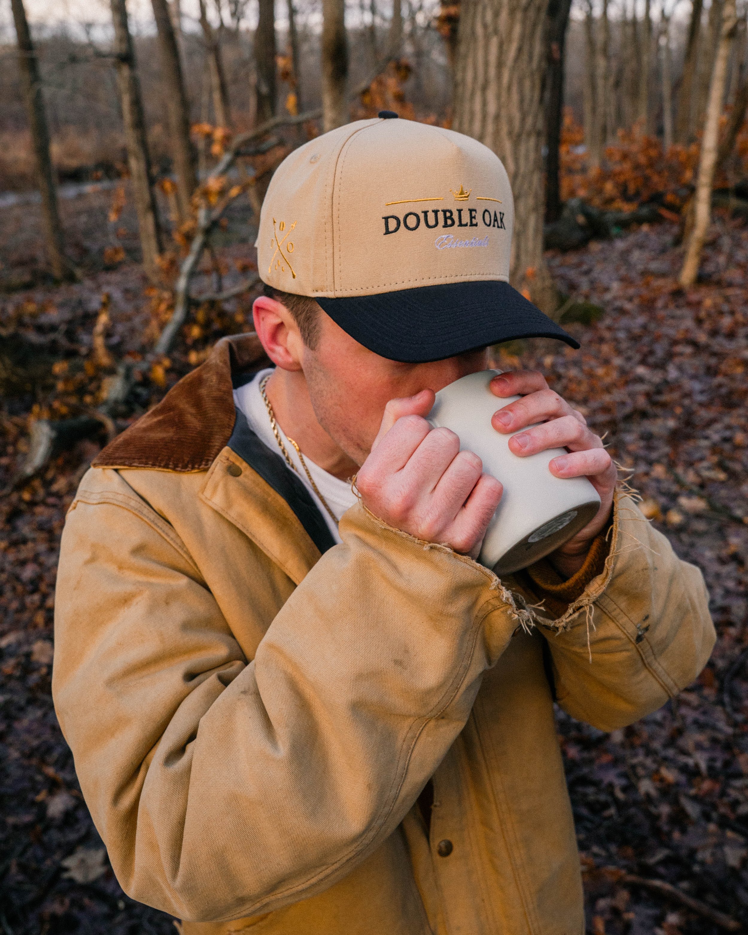 A person in a tan and black cap labeled 'Double Oak' drinks from a white mug in a wooded outdoor setting with fallen leaves.