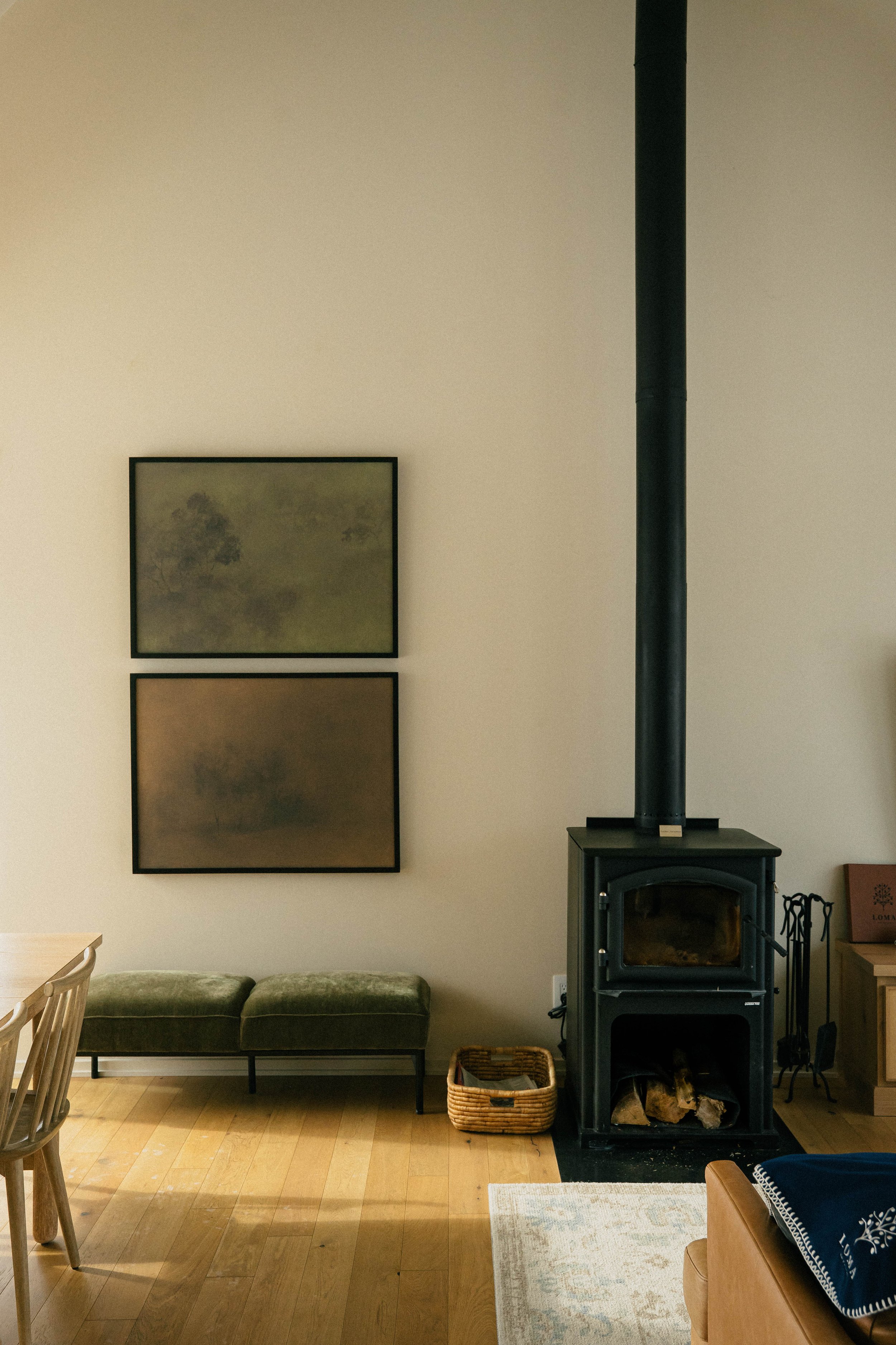 Living room with wood floor, two framed art pieces on wall, black wood stove with logs inside, small wicker basket, green upholstered bench, and part of a wooden table and chairs.