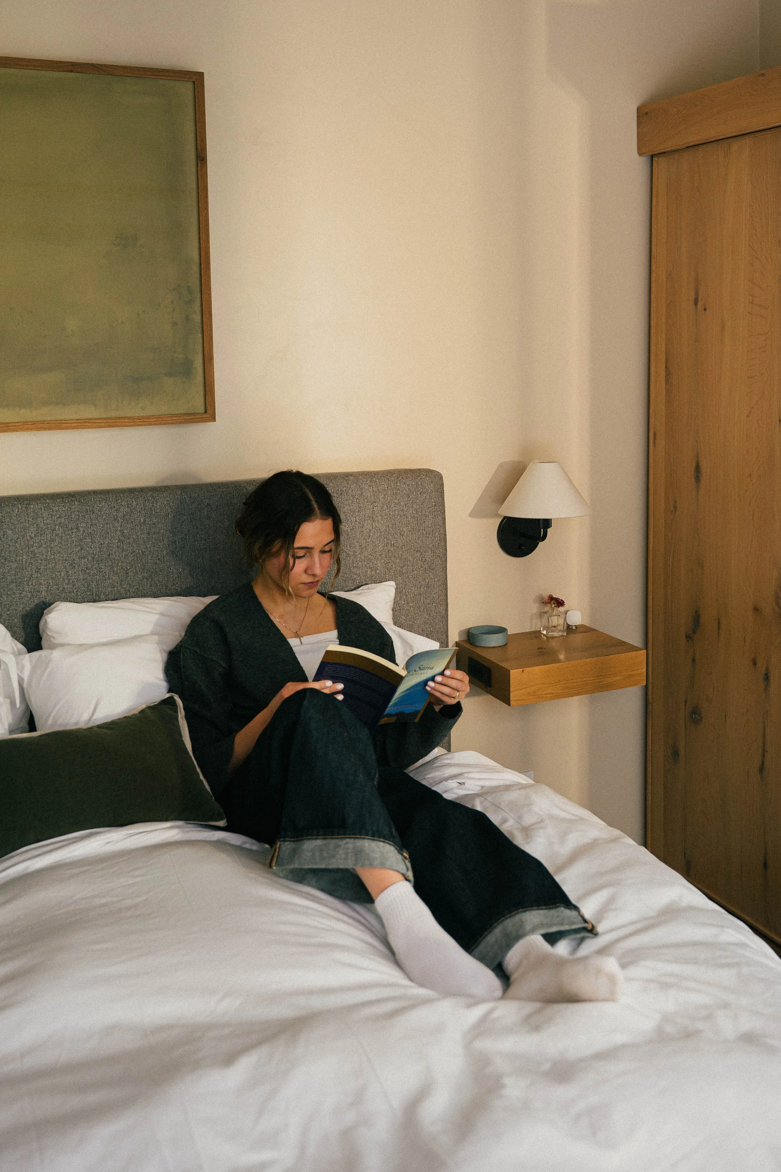A woman with dark hair and light skin tones sitting up in bed, reading a book. She wears a black top, gray sweatpants, and white socks. The bed has white linens and dark pillows. A nightstand with a small lamp, a vase with dried flowers, and a cup is