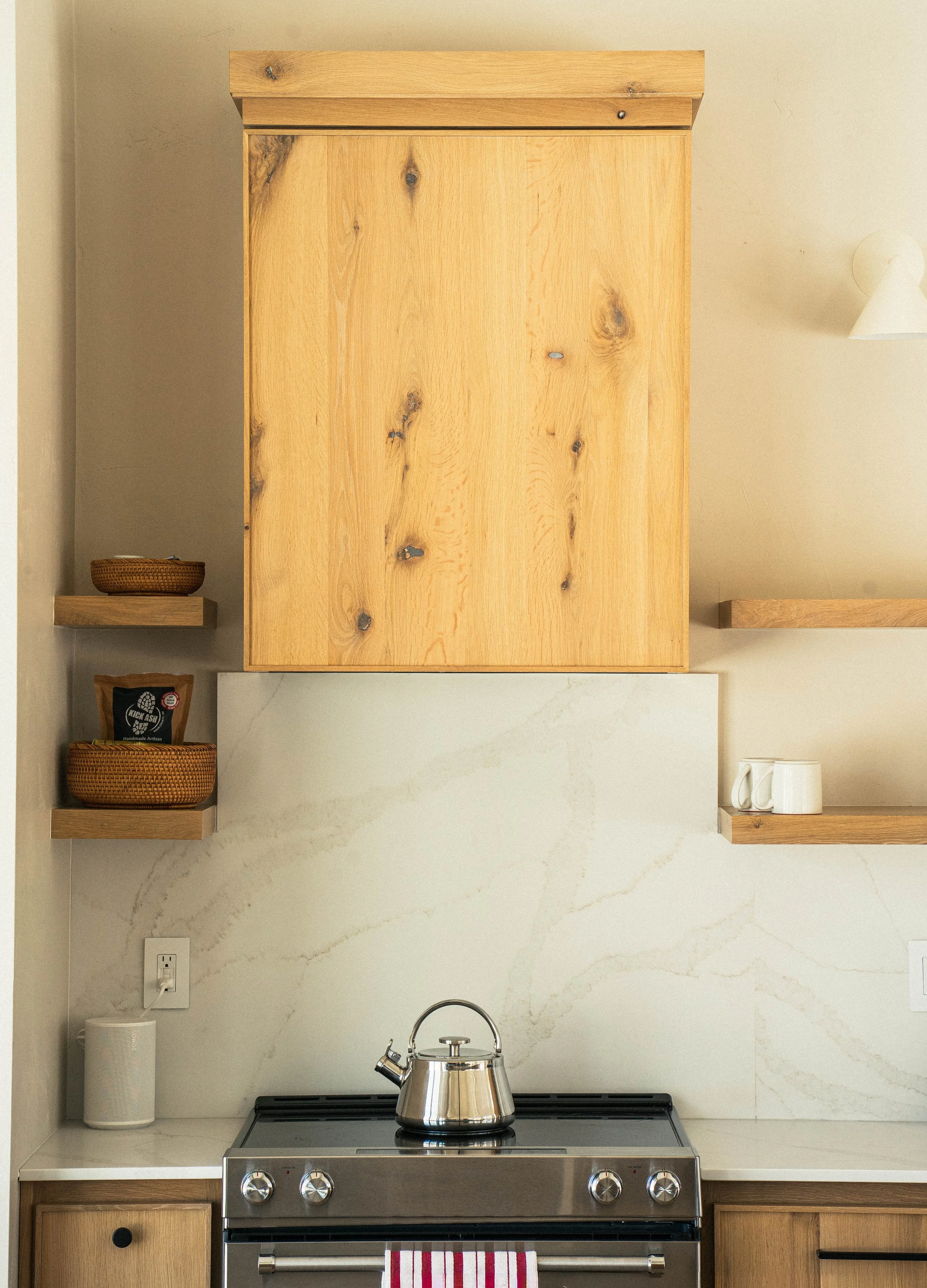 A kitchen stove with a stainless steel kettle on top of it, surrounded by beige marble countertops, with wooden cabinets above and below, and a white wall with a power outlet, a paper towel holder with a paper towel roll, and small shelves holding a 