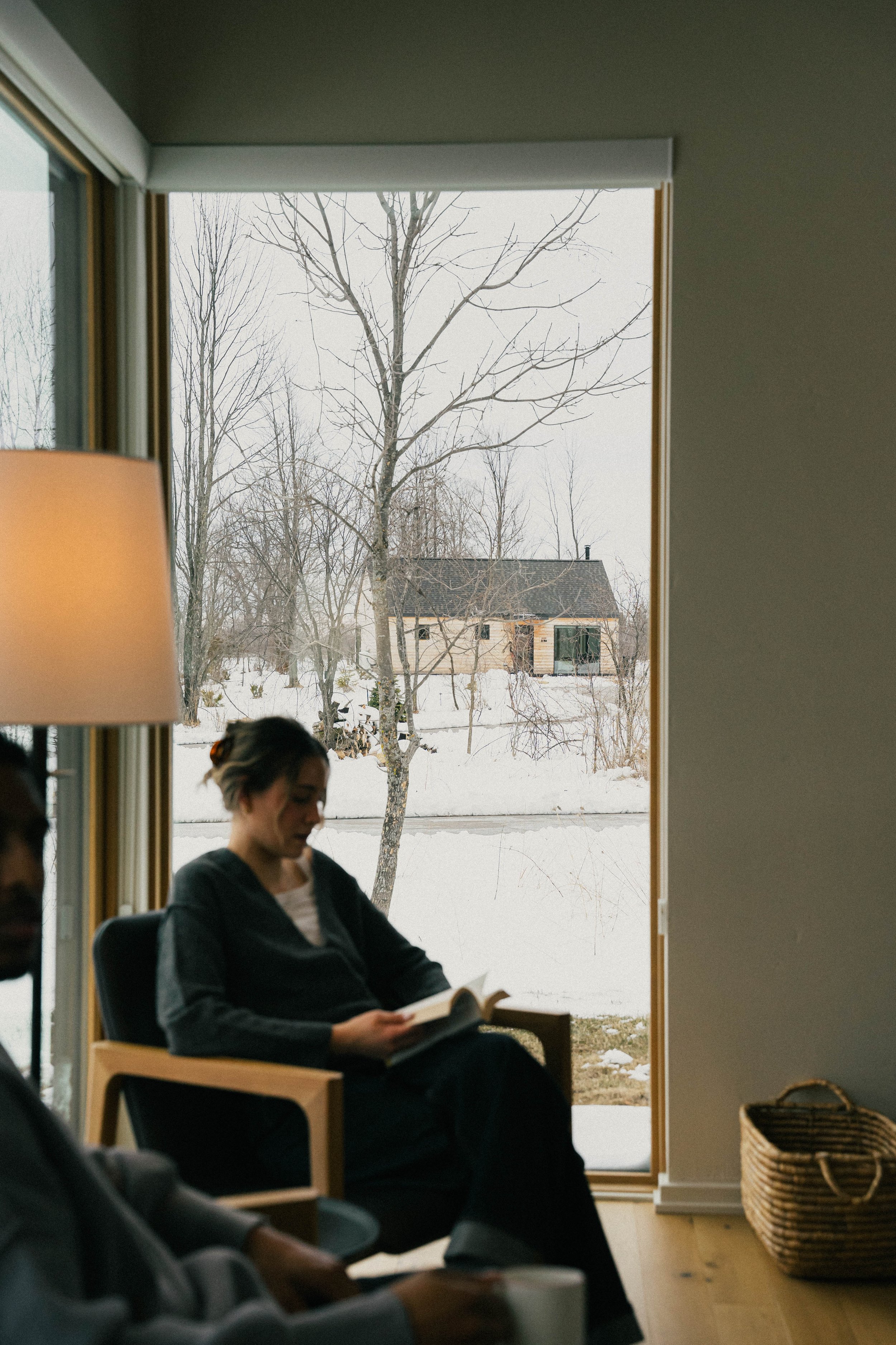 A woman sitting indoors reading a book, with a large window behind her showing a snowy yard and leafless trees outside.
