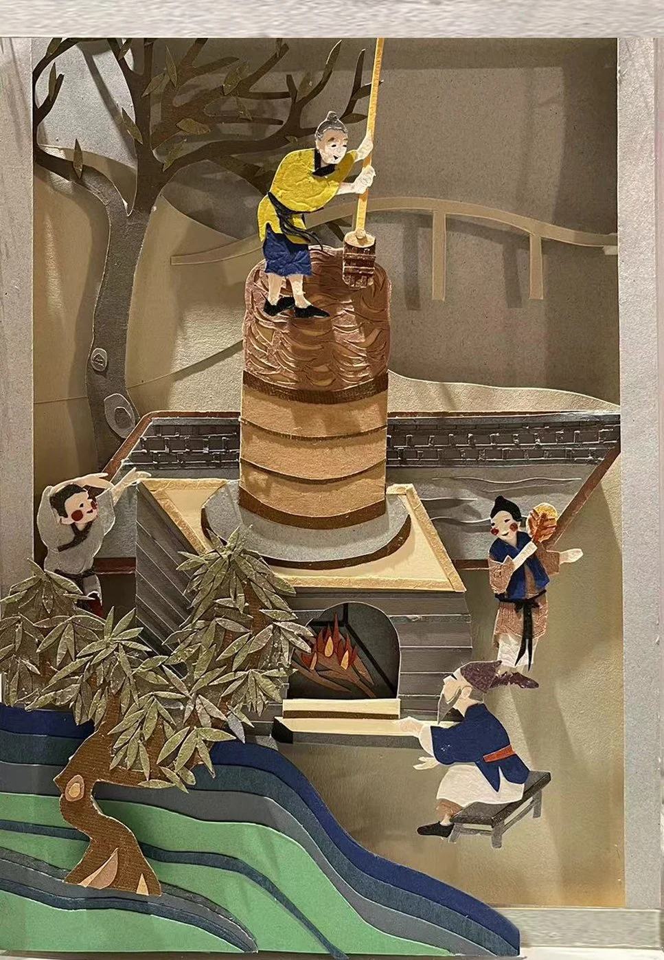 A paper craft scene of children around a campfire with a large brown tree, a person roasting marshmallows, and others playing or watching near the fire, set on a layered landscape.