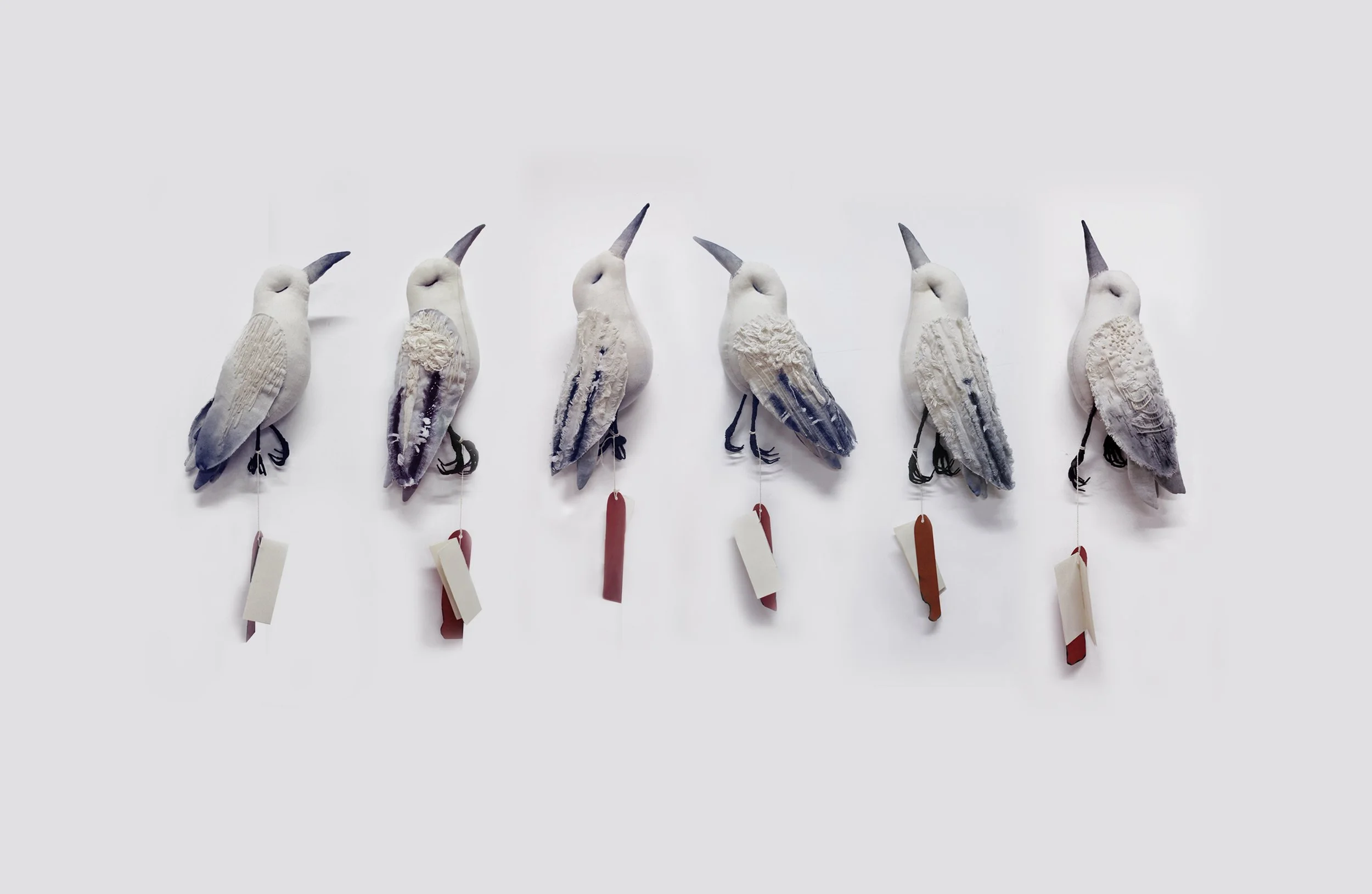 Wall display of six decorative bird sculptures designed to resemble songbirds, each with a tag hanging from their beak.