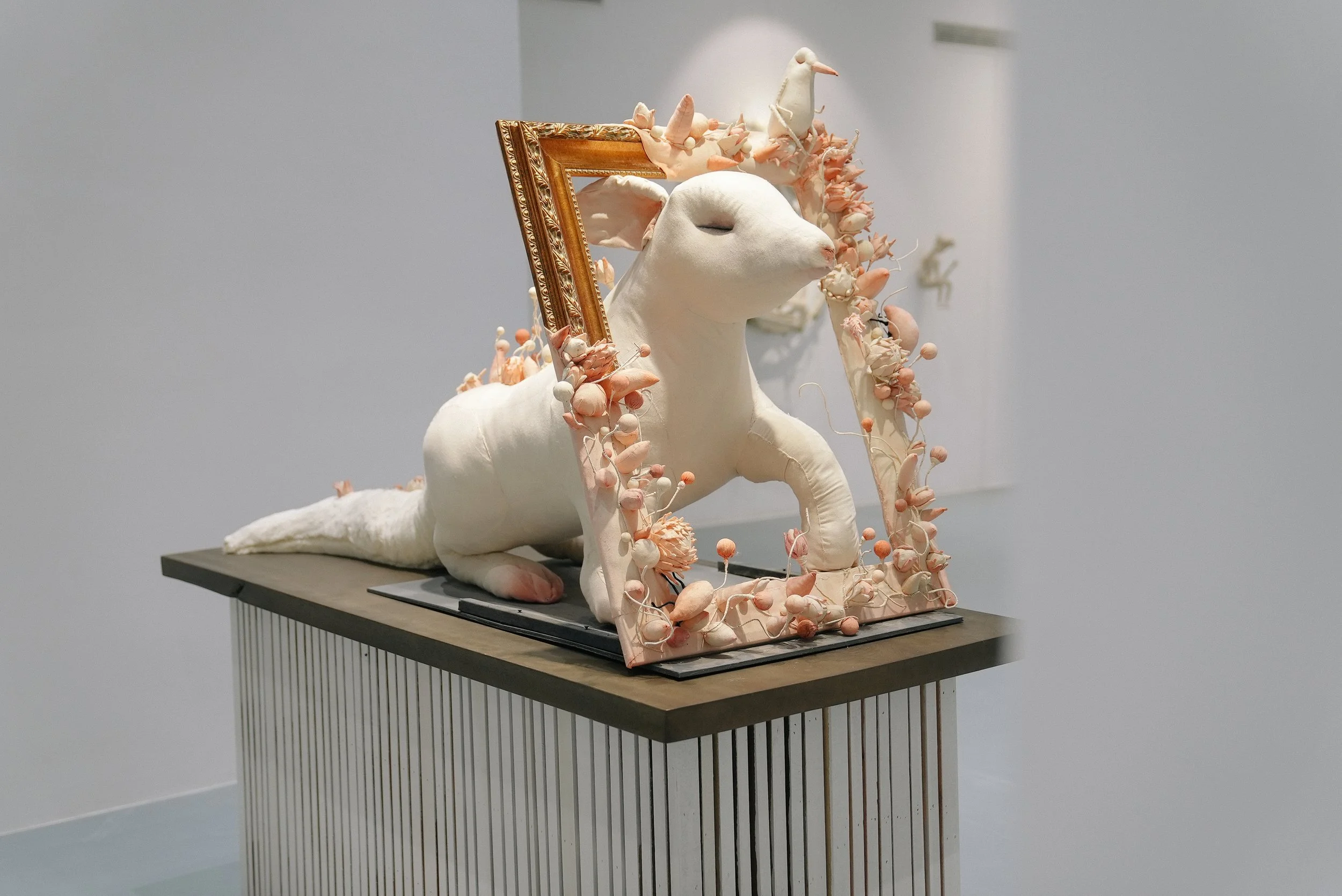 An artistic sculpture of a white dog lying on a table, adorned with pink and peach floral decorations and surrounded by a pink ornate frame.