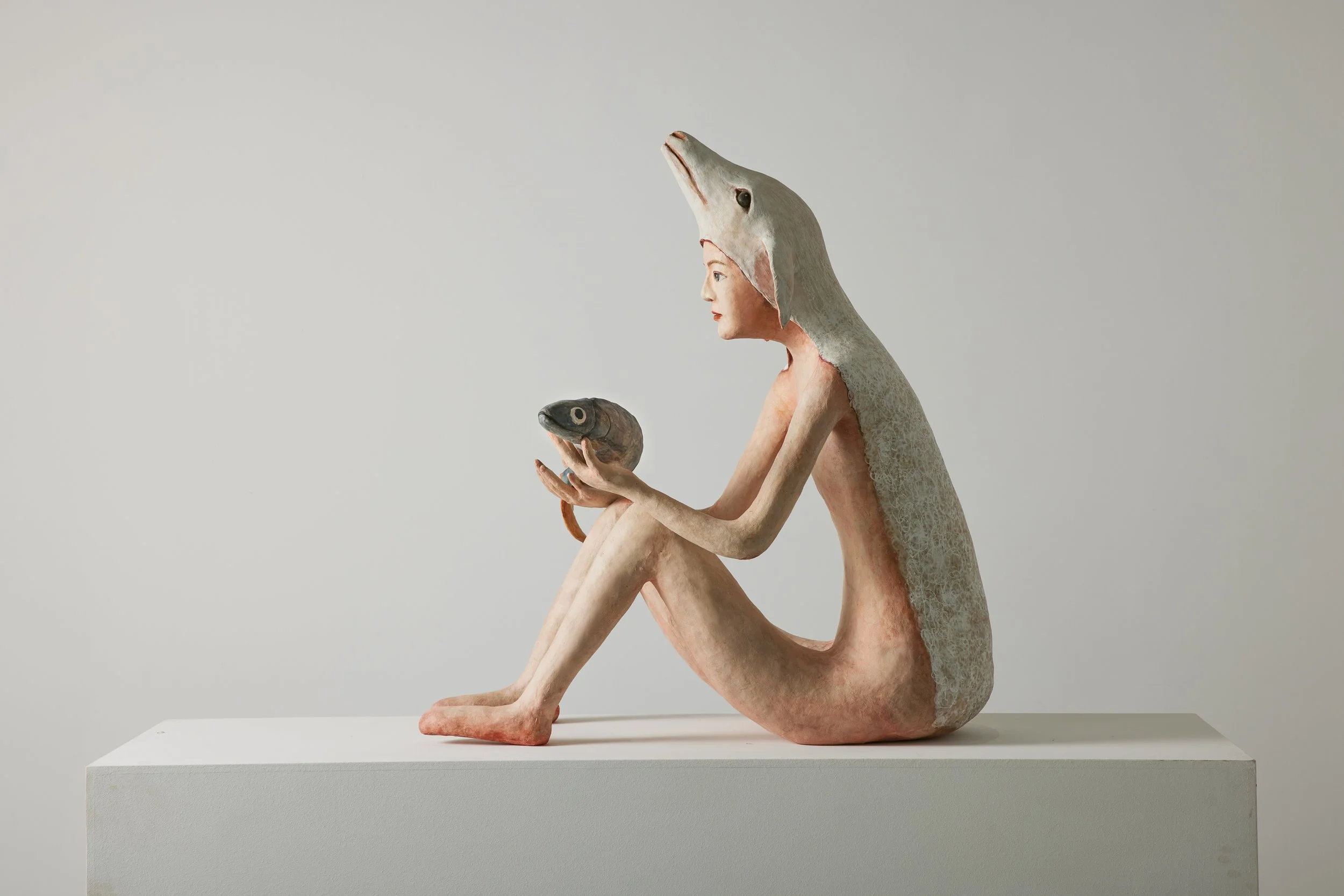 a sculpture of a seated character with a human face, wearing a whale costume, holding a fish, on a white pedestal against a plain background.