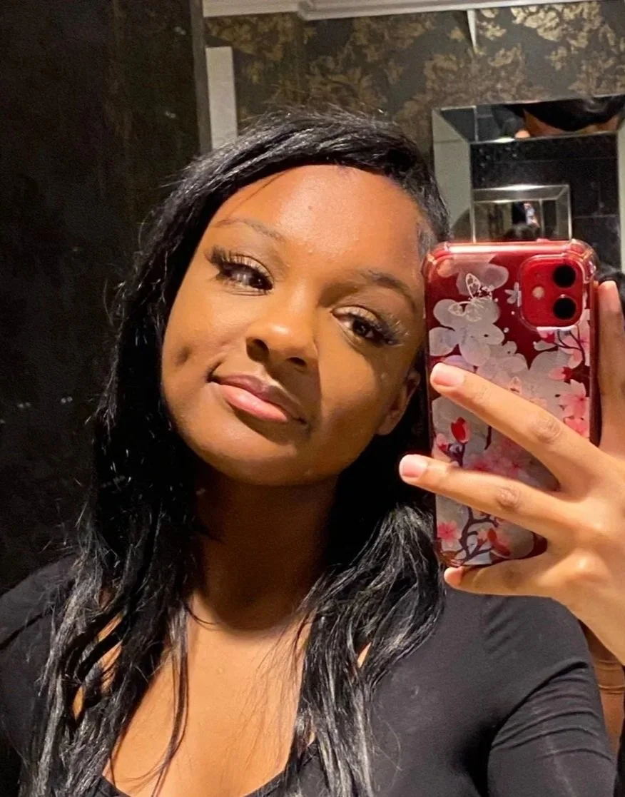 A woman with dark hair and makeup taking a selfie in a mirror with her phone, which has a floral patterned case.