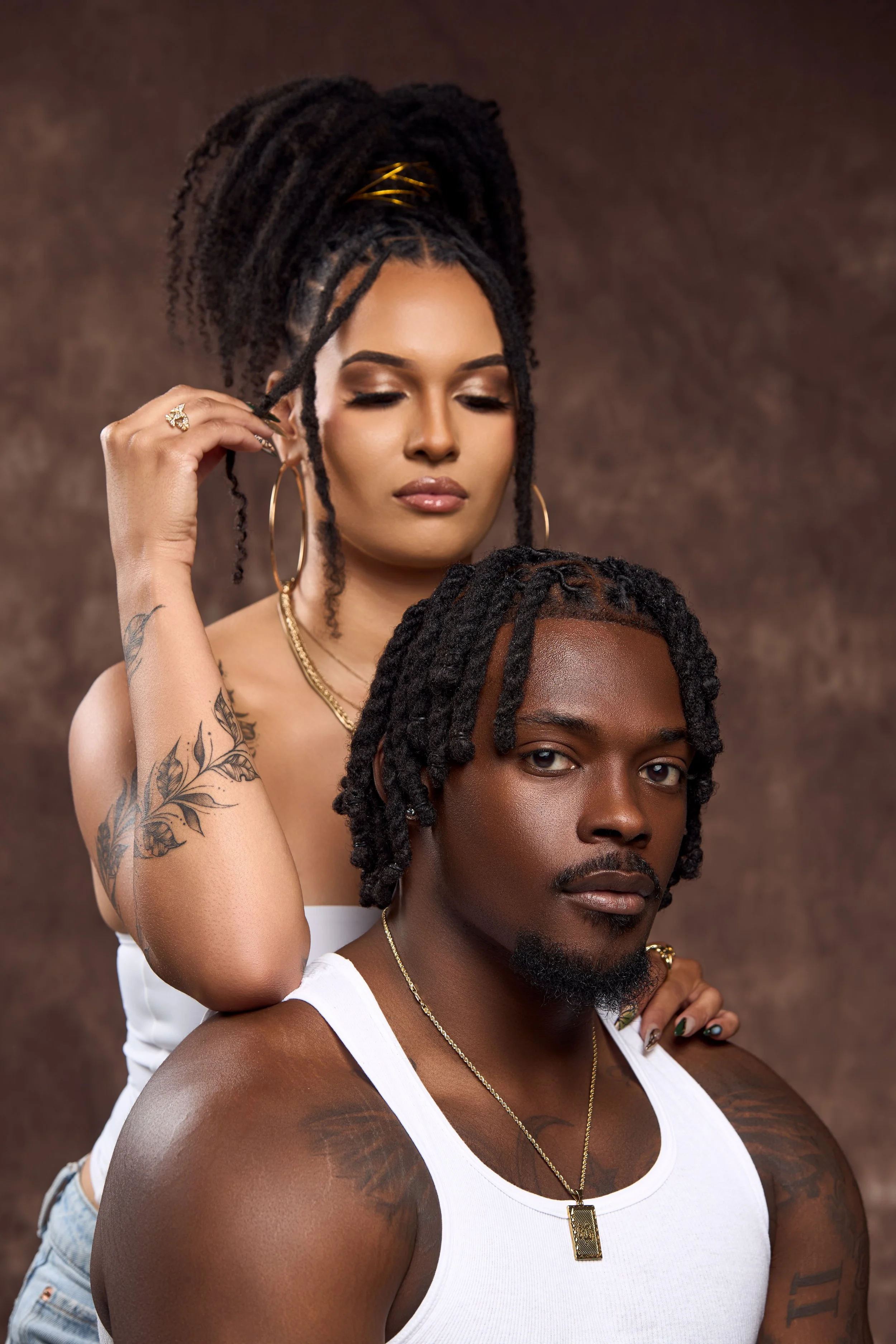 A woman with dreadlocks and large hoop earrings styling a man with dreadlocks and a white tank top, both posing against a brown background.