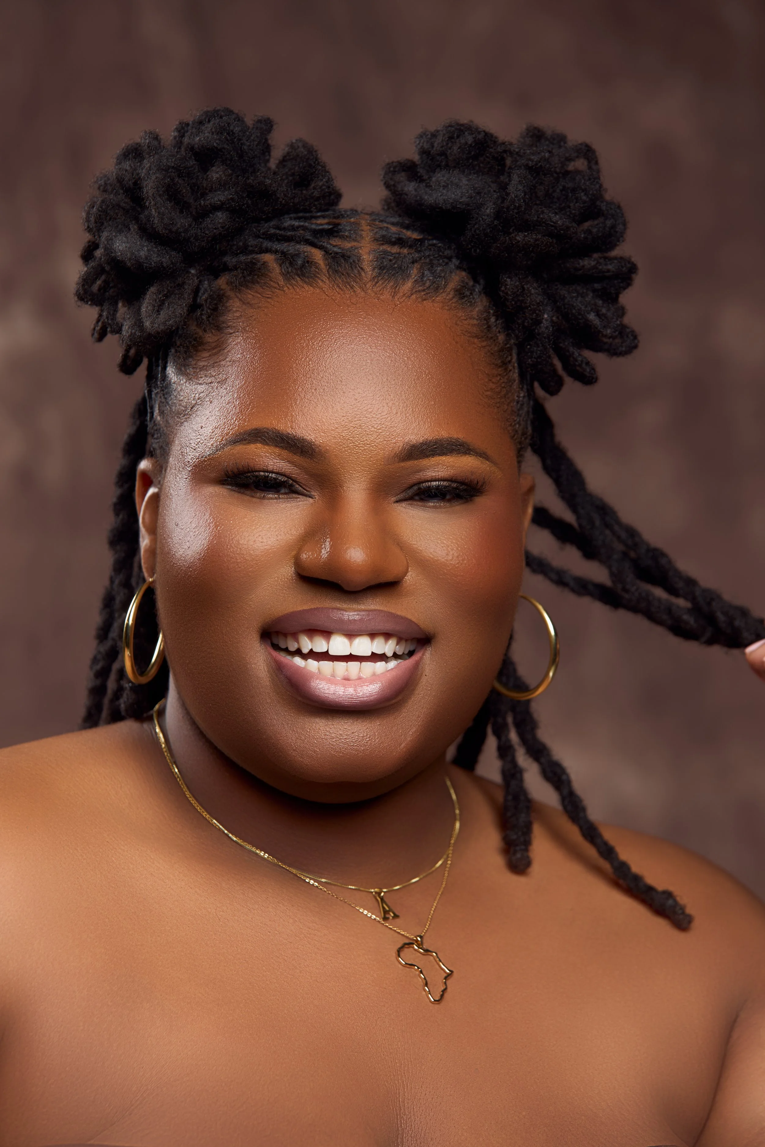 A smiling woman with dark skin and dreadlocks, wearing gold hoop earrings and layered gold necklaces, one with an Africa-shaped pendant, posing against a brown and beige background.