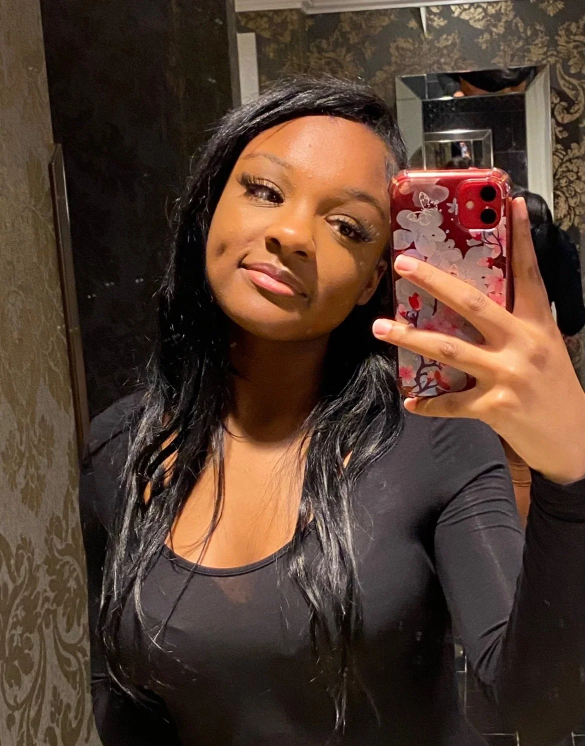 A woman taking a mirror selfie with a decorative phone case in a bathroom with dark wallpaper.