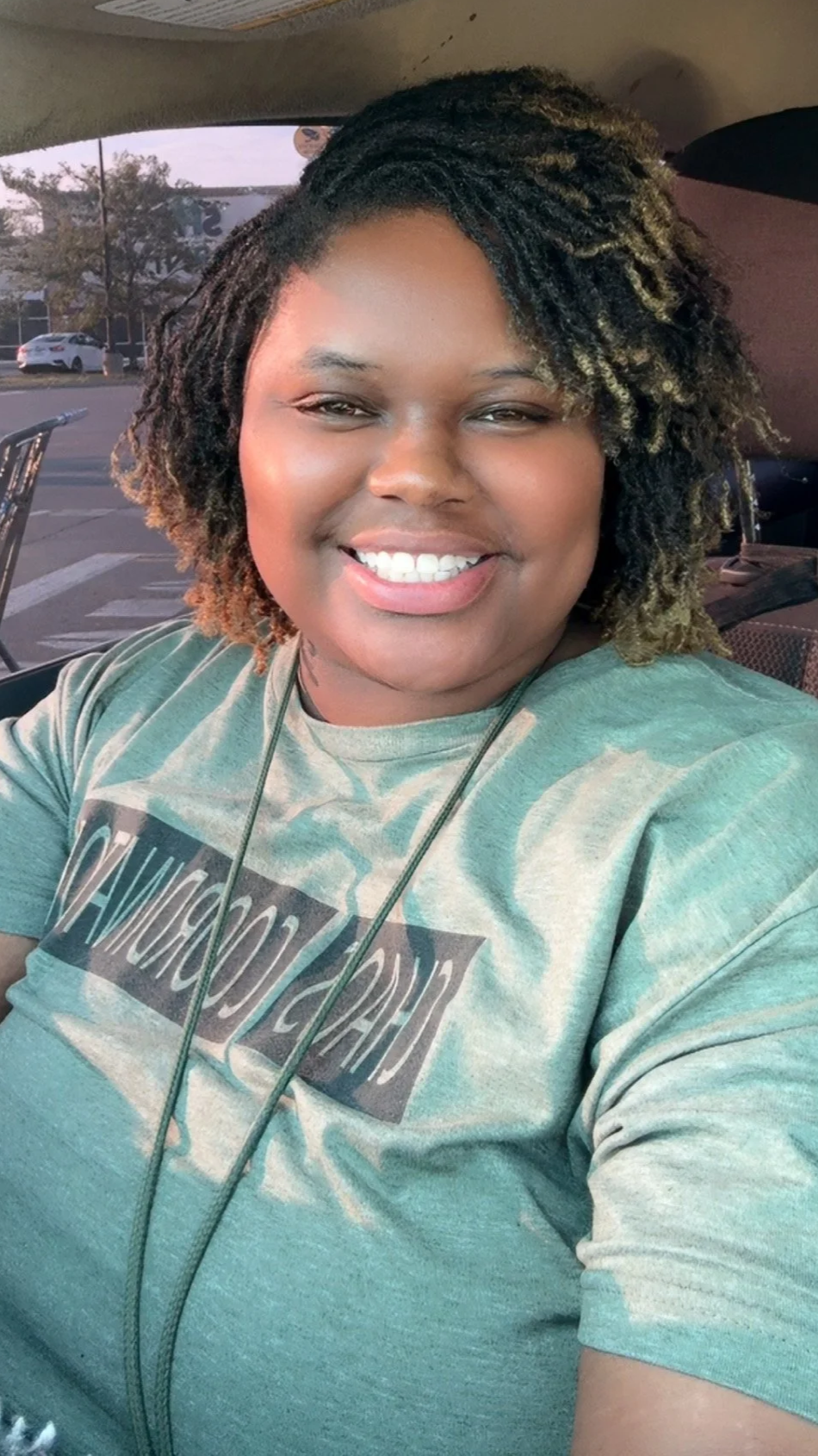 A woman smiling and taking a selfie inside a vehicle with a parking lot and trees visible through the window behind her.