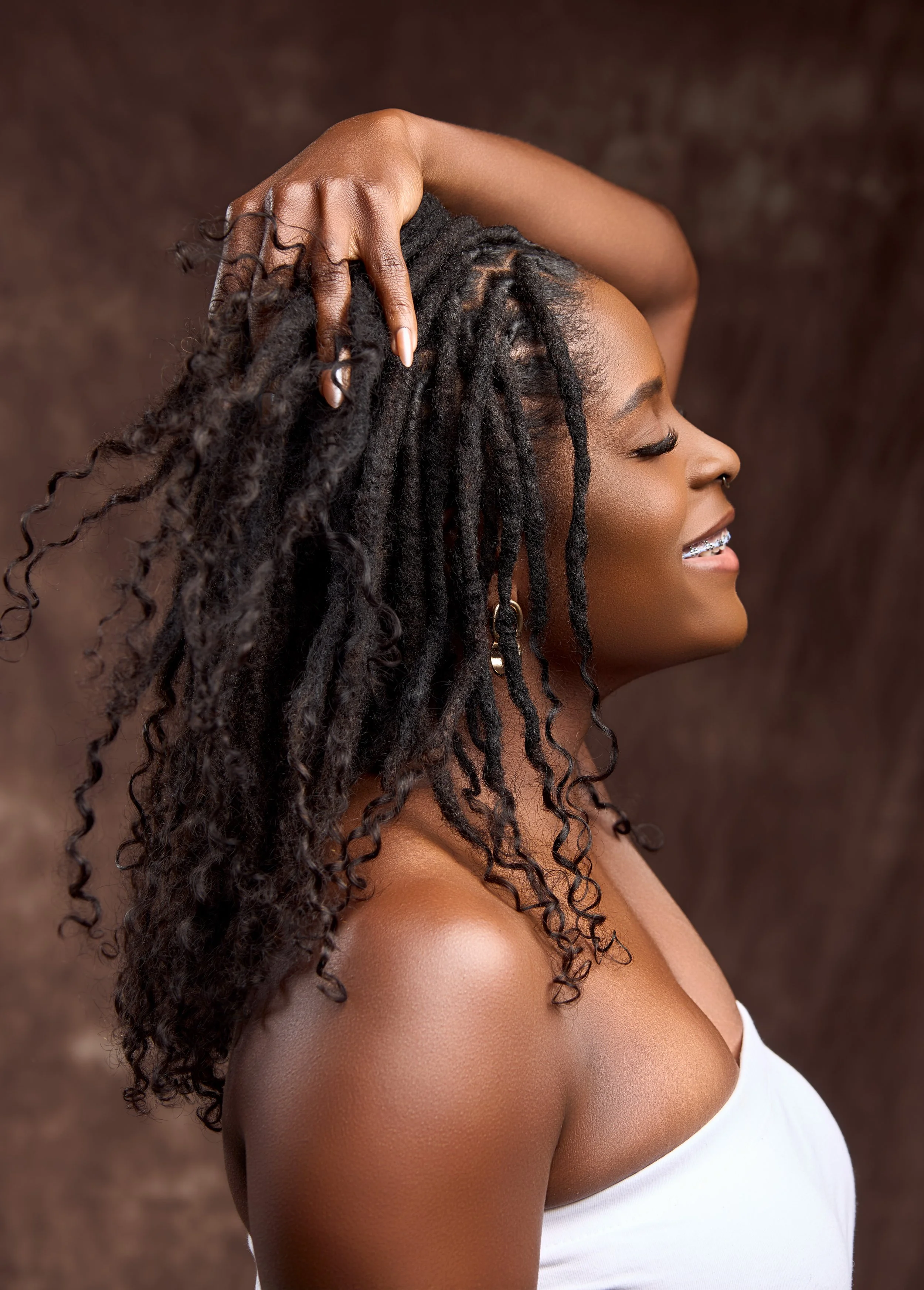 A woman with natural dreadlocked hair, wearing a white top, smiling with closed eyes in profile view.