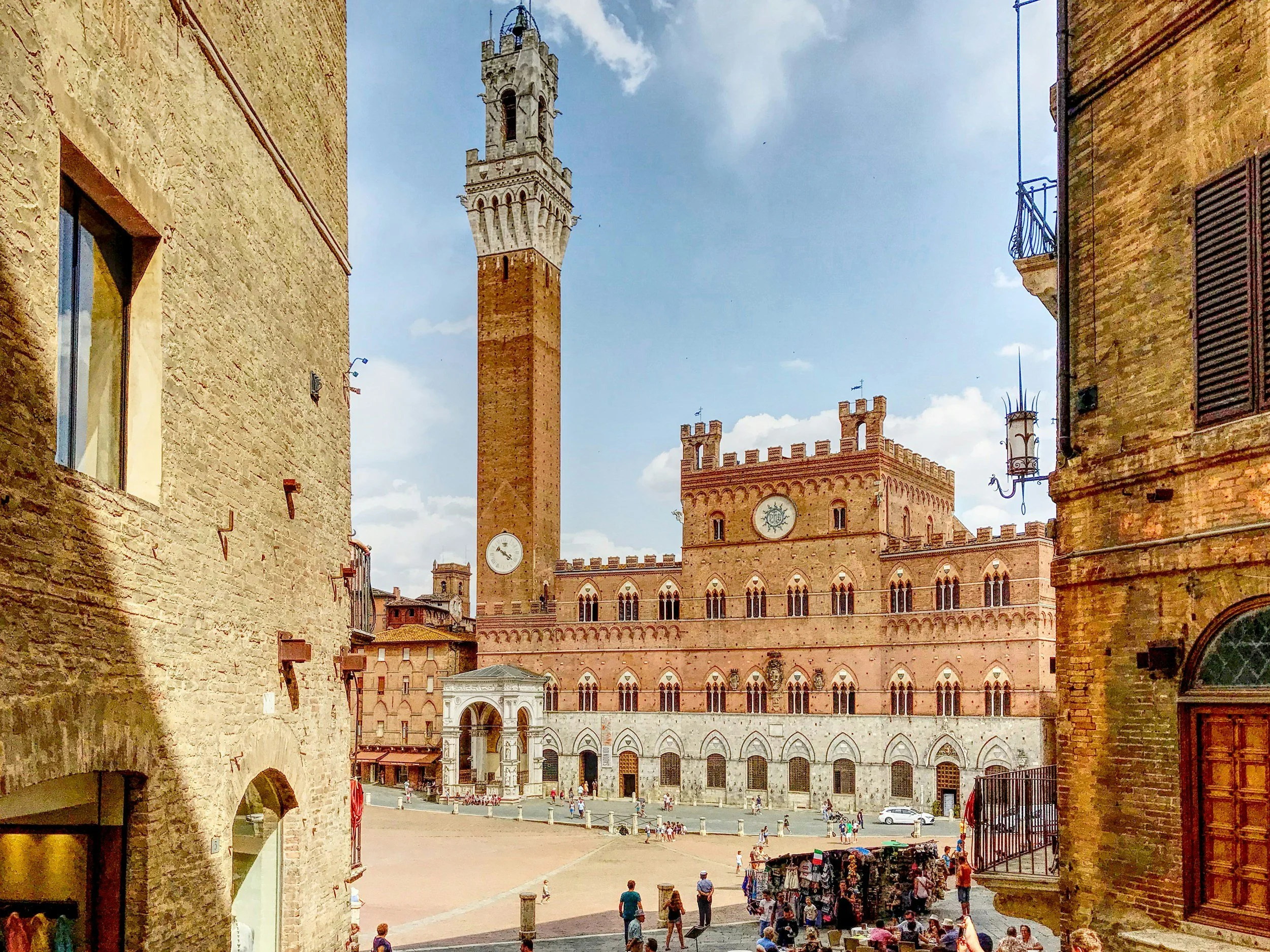 People walking and shopping in a historic square with brick buildings, a tall clock tower, and a castle-like structure with crenellations.