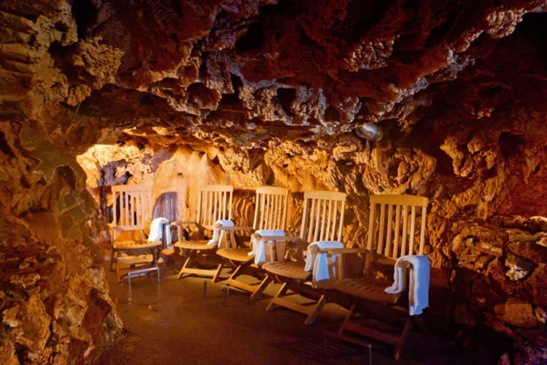 A cave with textured, rocky walls illuminated with warm orange lighting. Six wooden chairs with armrests are lined up along one side, each draped with a white towel.