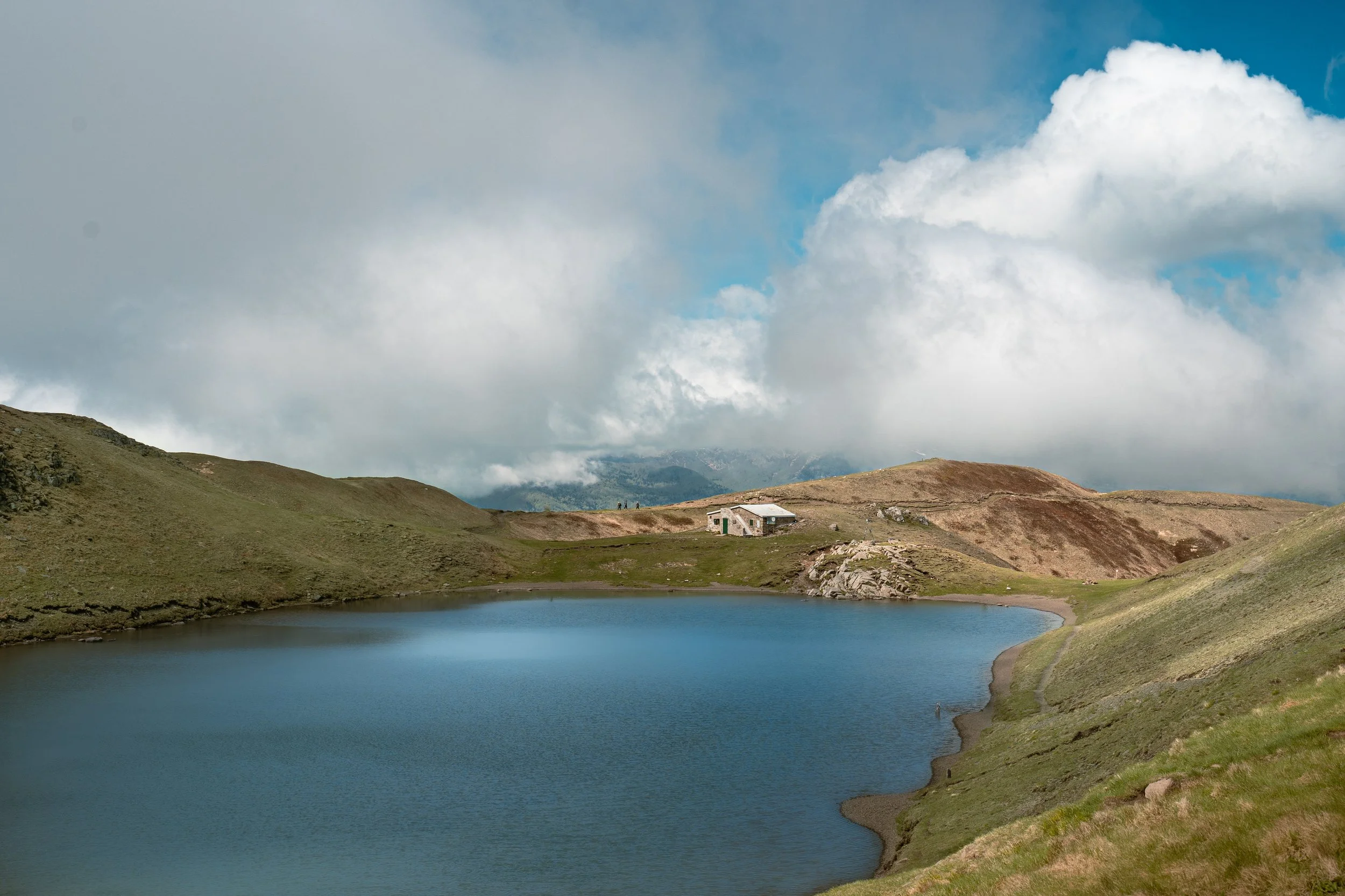 A scenic mountain landscape with a small lake, rolling green hills, and a rustic house, under a partly cloudy sky.
