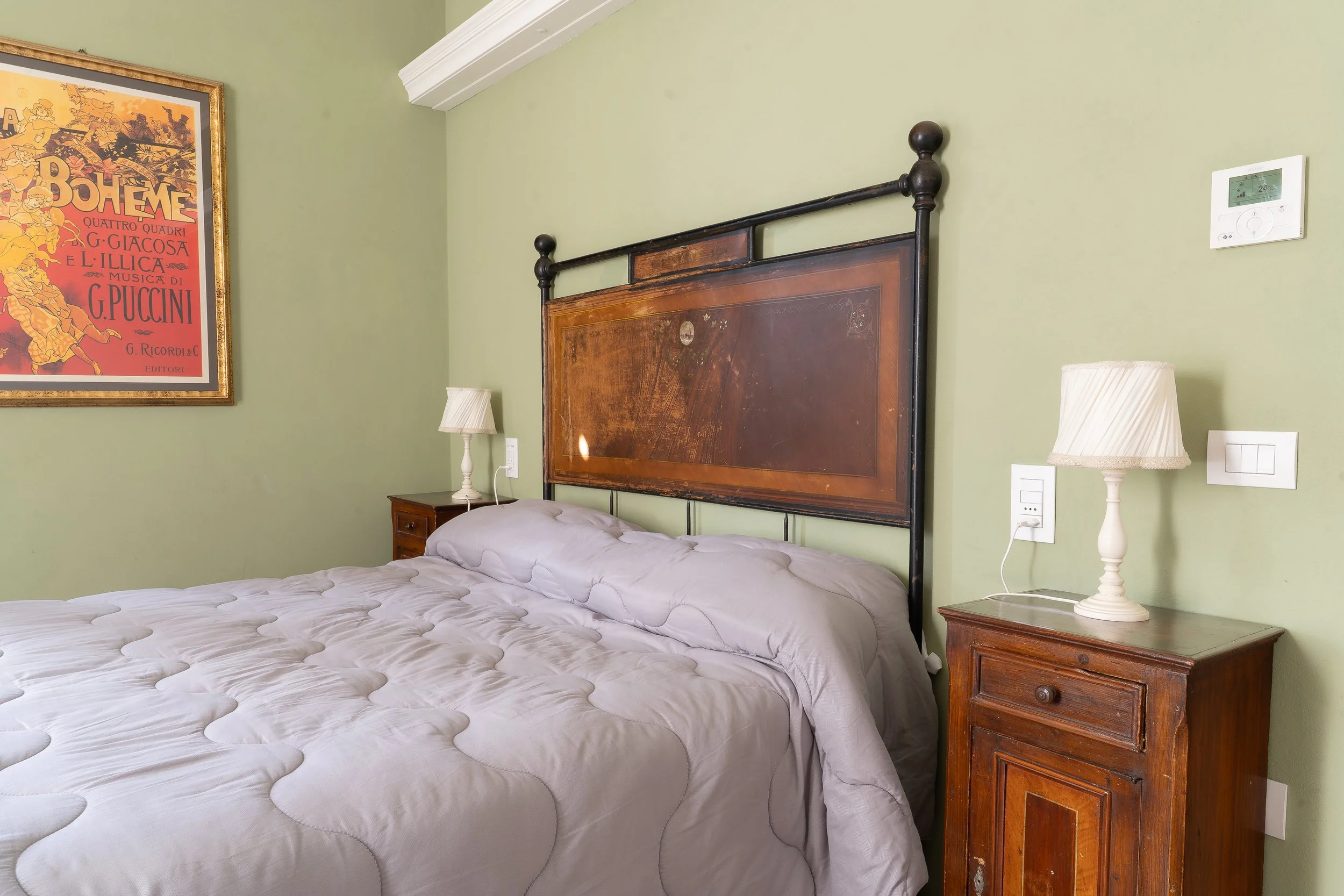 A bedroom with a green wall, a wooden headboard, and two matching wooden nightstands with white lamps, a framed vintage poster, and a thermostat.