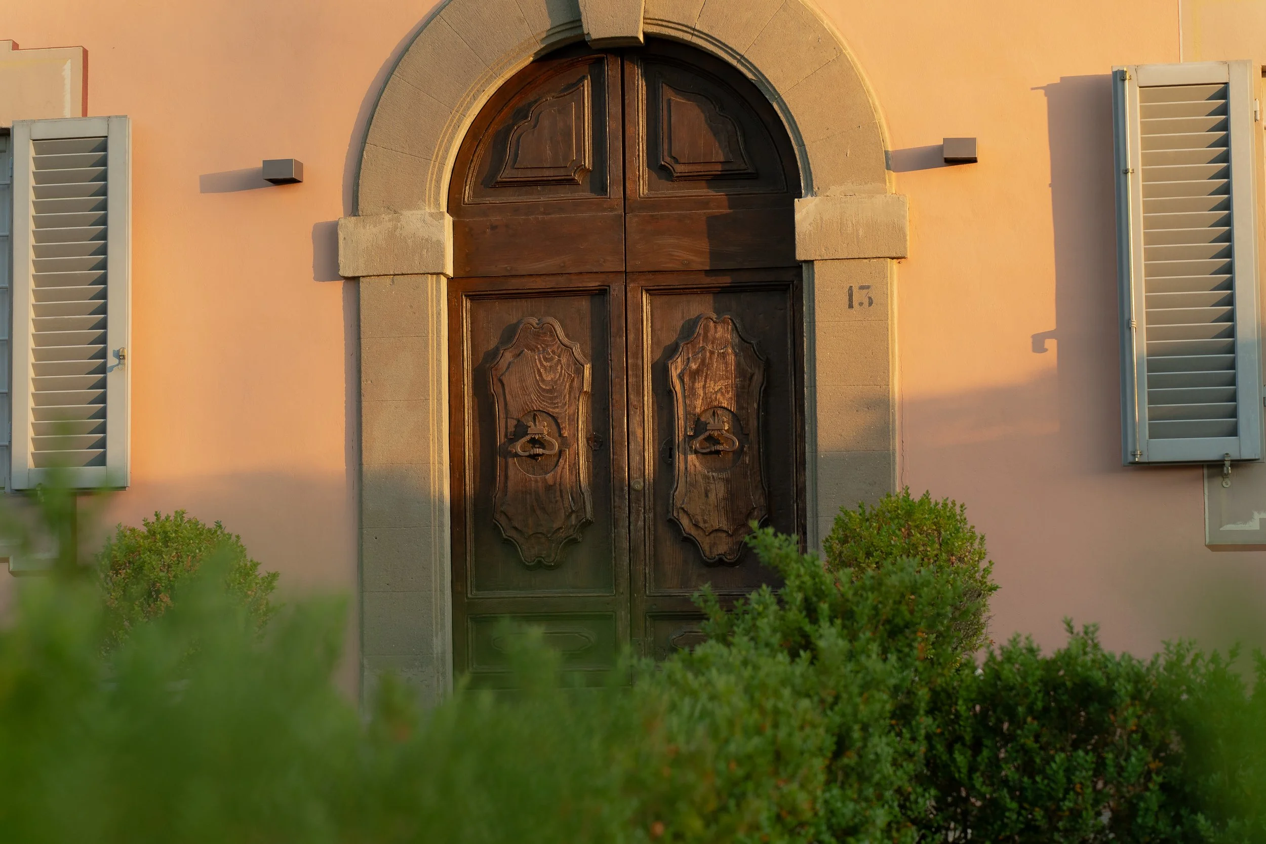 Wooden front door with decorative panels, set in a stone archway, on a peach-colored building with white shutters and green bushes in the foreground.
