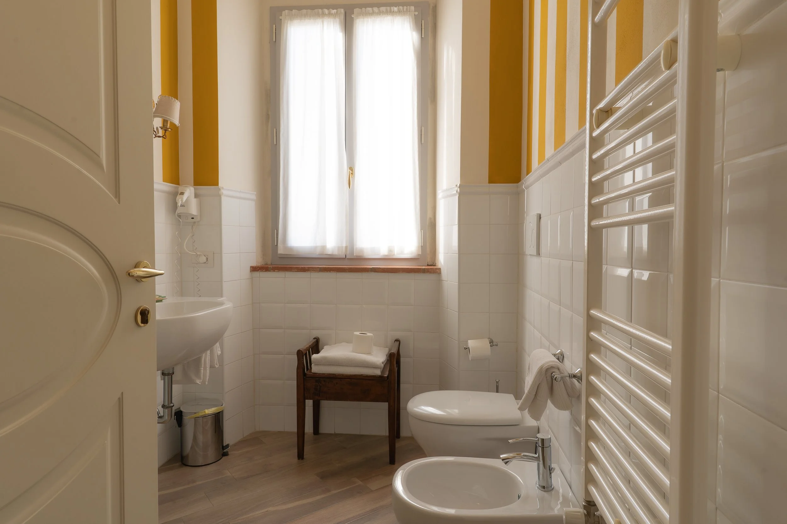 A small bathroom with white tiles and yellow painted walls. Features include a small sink, a toilet, a bidet, a wall-mounted towel rail, a towel, a window with white curtains, and a wooden tray with folded towels and a roll of toilet paper. There is 