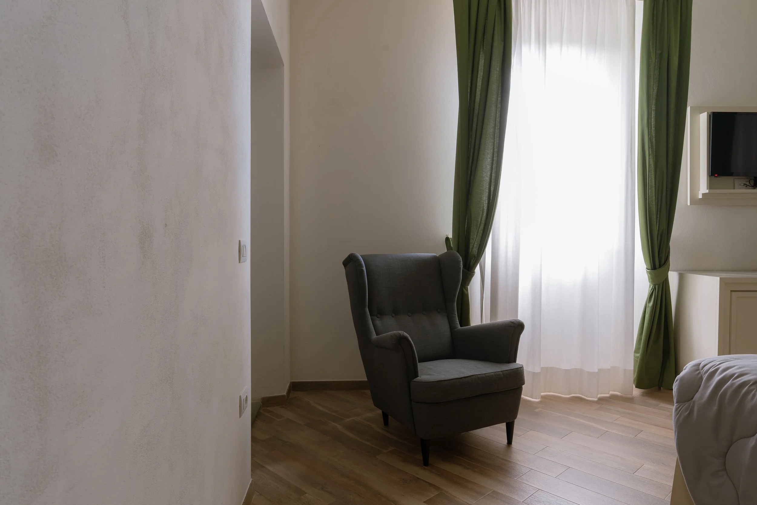 A cozy corner with a high-backed gray armchair next to a window with green curtains, sheer white curtains behind, and a wall-mounted TV on a shelf in a bright room with wooden flooring.