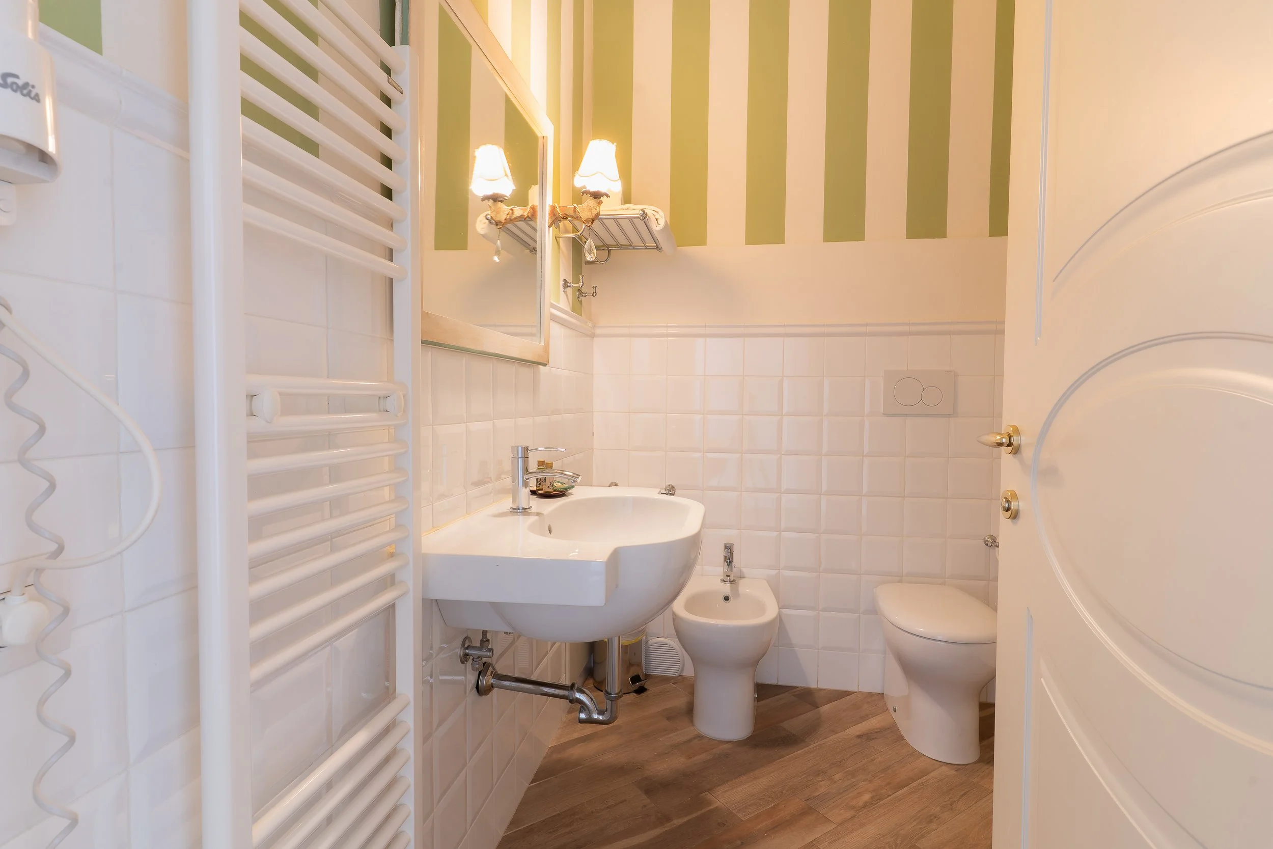 Bathroom with white tiled walls, a white sink, a bidet, a toilet, and a wall-mounted mirror with two lamps. There is a white towel radiator on the left and a wooden floor.