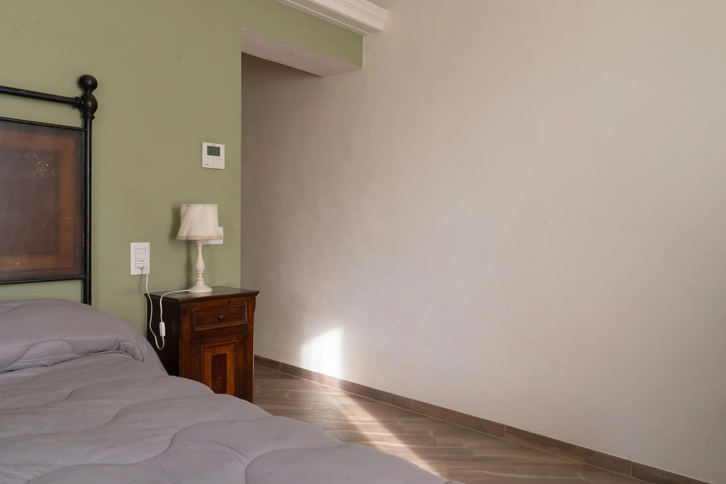 A bedroom corner with a bed, wooden nightstand with a lamp, a wall-mounted thermostat, and a light switch, with sunlight on the floor.