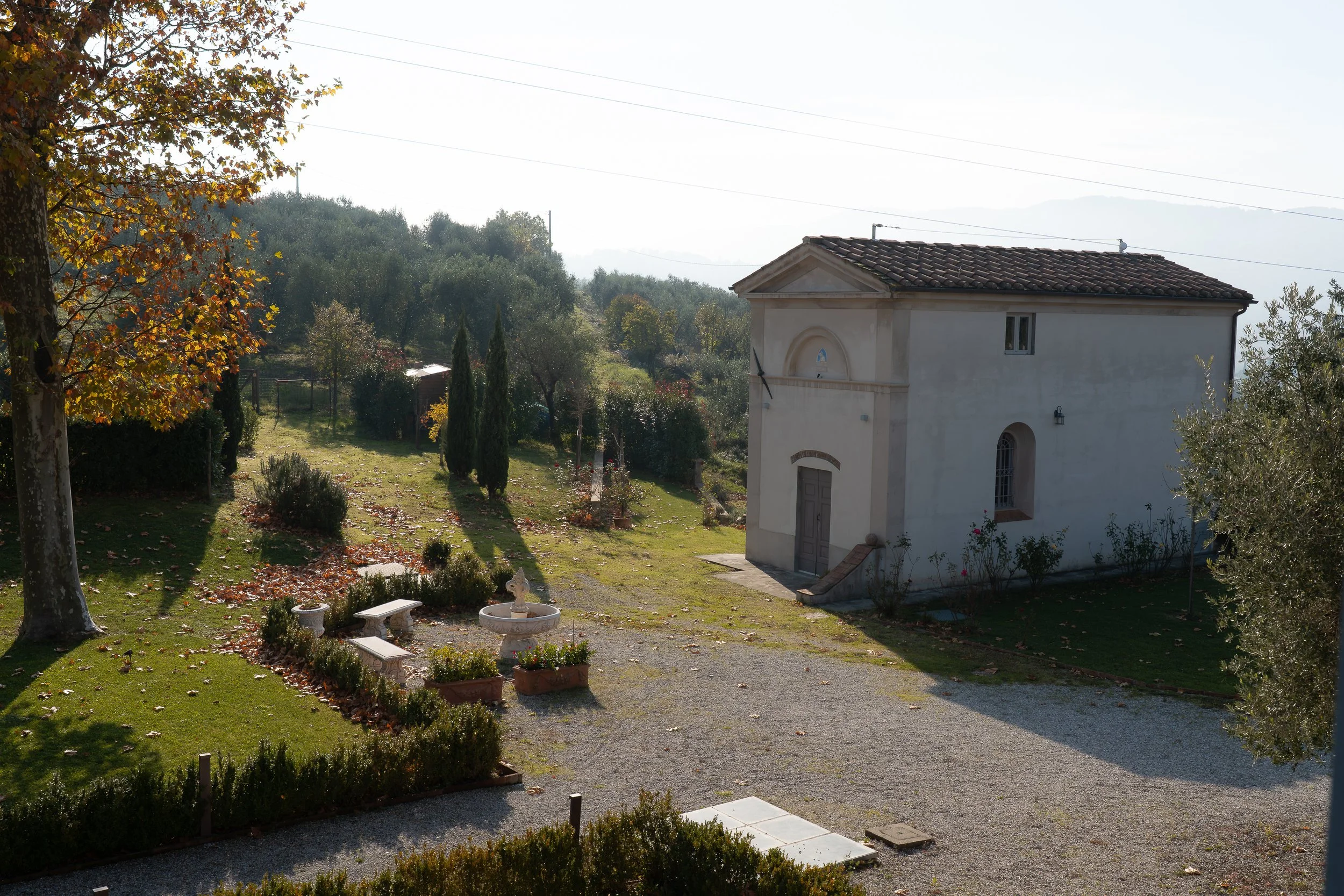 A small white church with a brown tiled roof, surrounded by a garden with trees, bushes, and a gravel pathway, during daytime with sunlight.