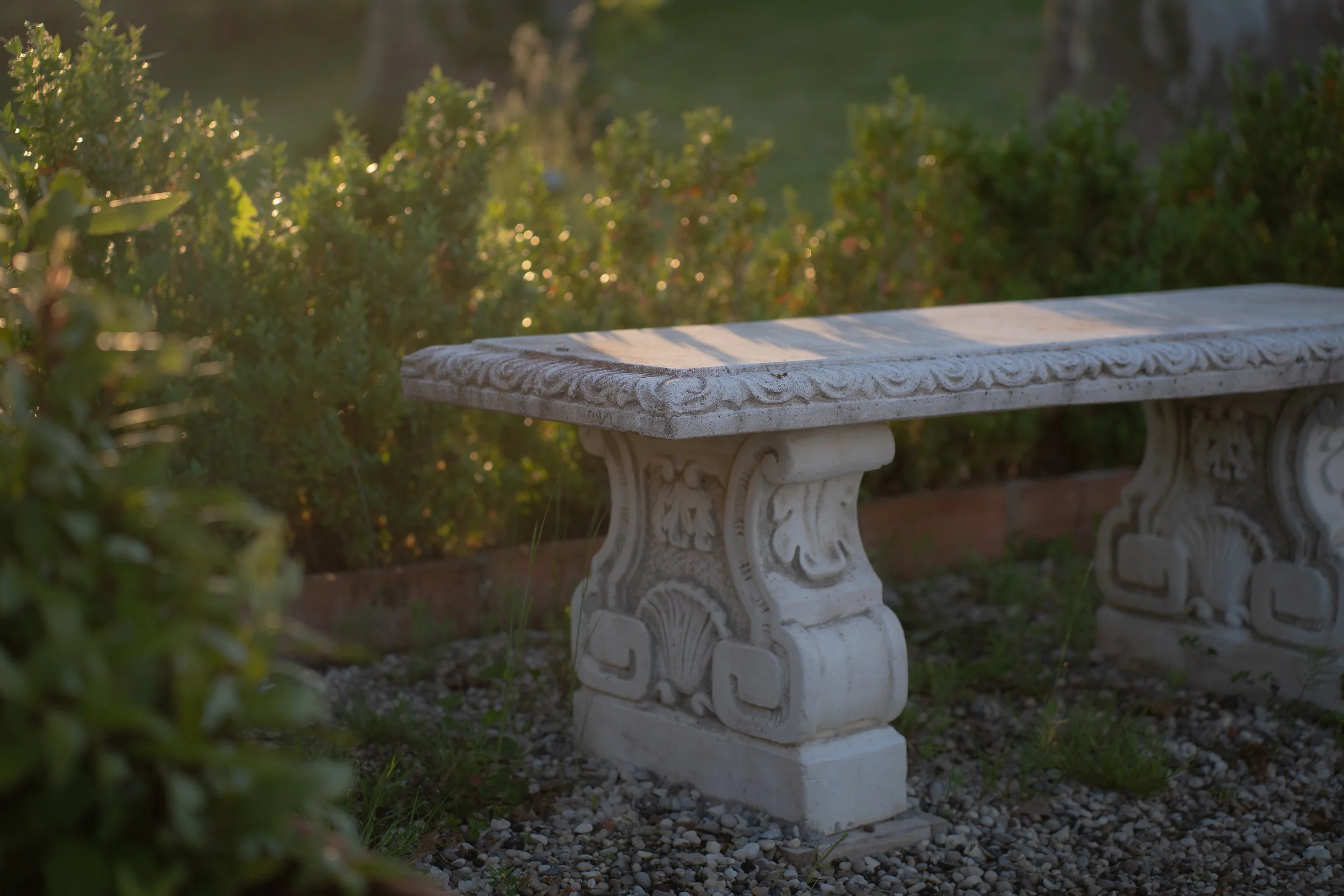 White stone decorative bench outdoors at sunset, surrounded by greenery.