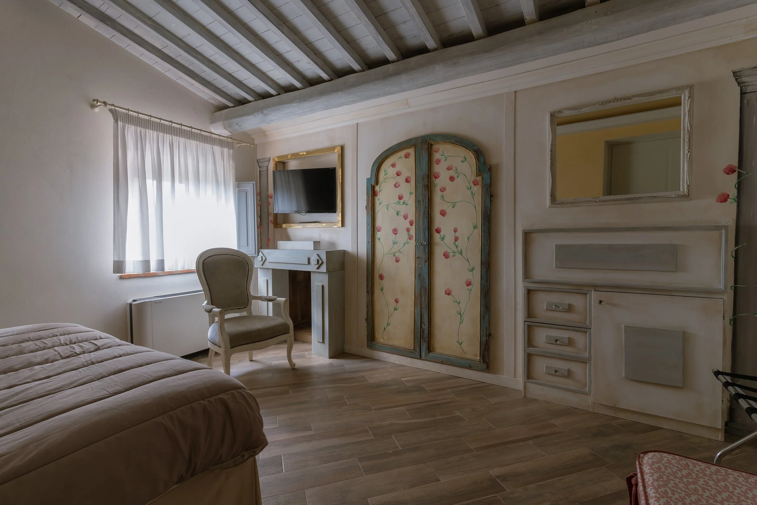 A bedroom with a bed, a window with white curtains, a beige armchair, a wall-mounted TV, an ornate wardrobe with floral design, a mirror, and a fireplace.