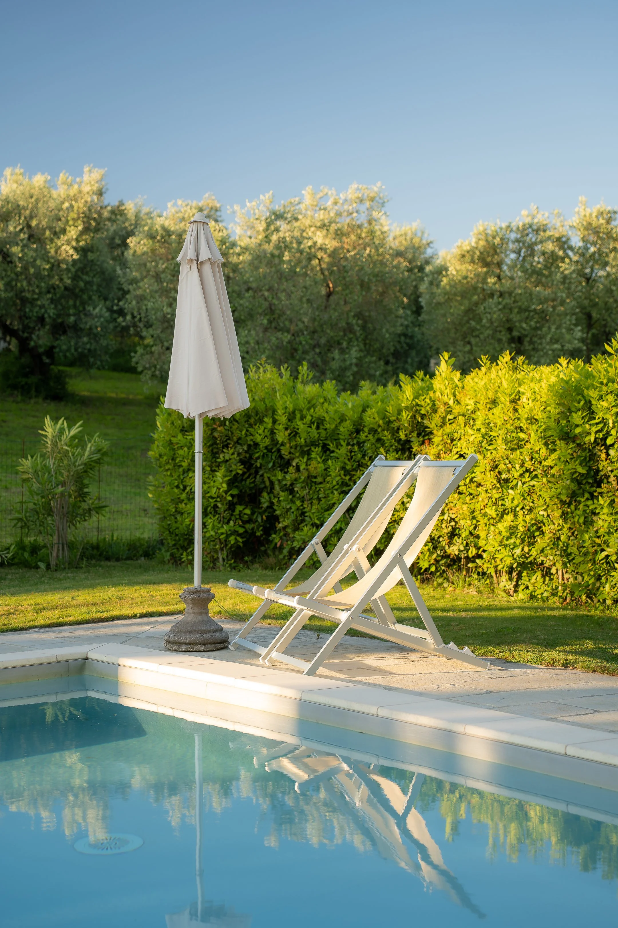 Two lounge chairs and a closed umbrella next to a swimming pool on a sunny day with trees in the background.