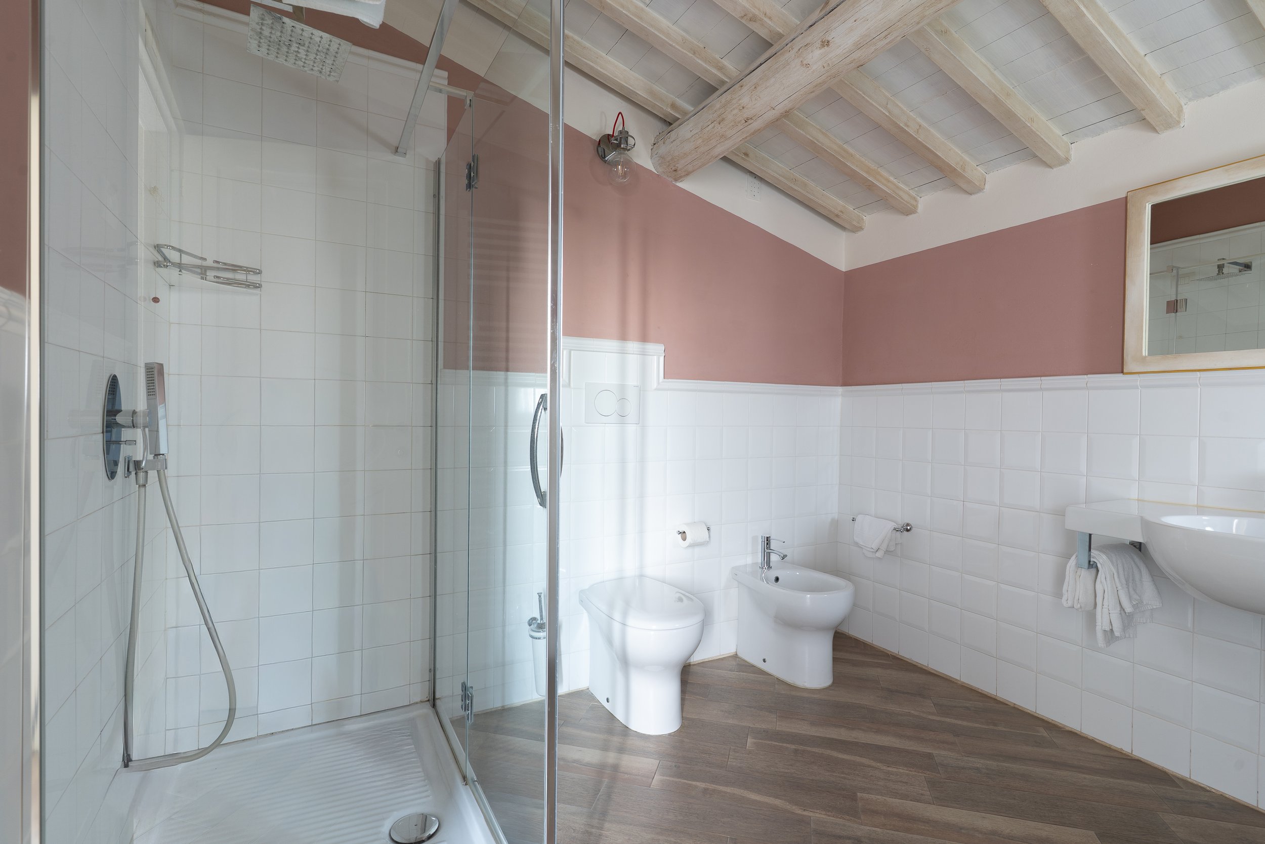 Bathroom with a walk-in shower, toilet, bidet, small sink, mirror, white tiled walls, pink upper wall, wooden ceiling beams, and wooden flooring.
