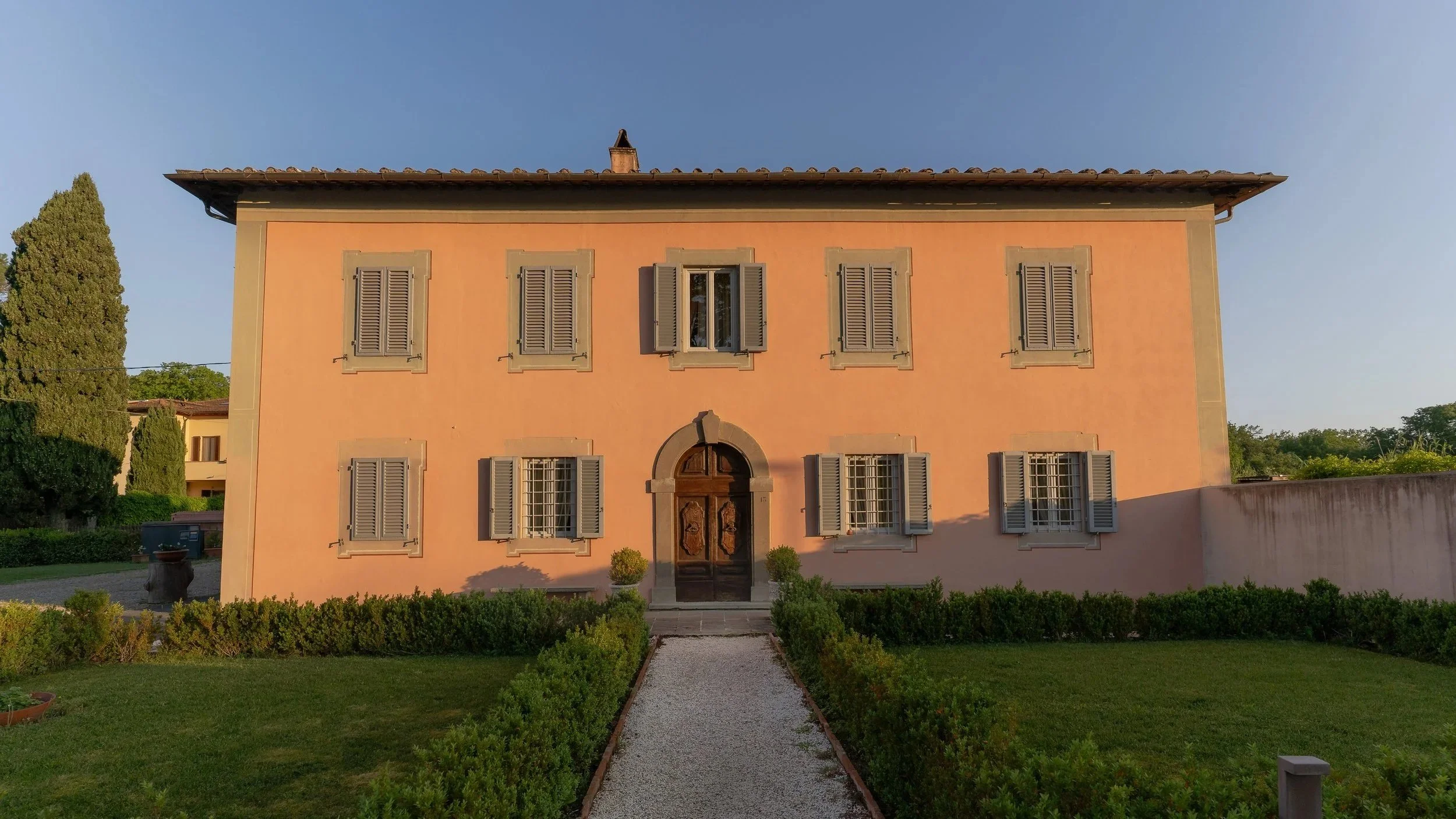 A two-story house with pink stucco walls, gray window shutters, and a dark wooden front door, surrounded by greenery and a gravel pathway.