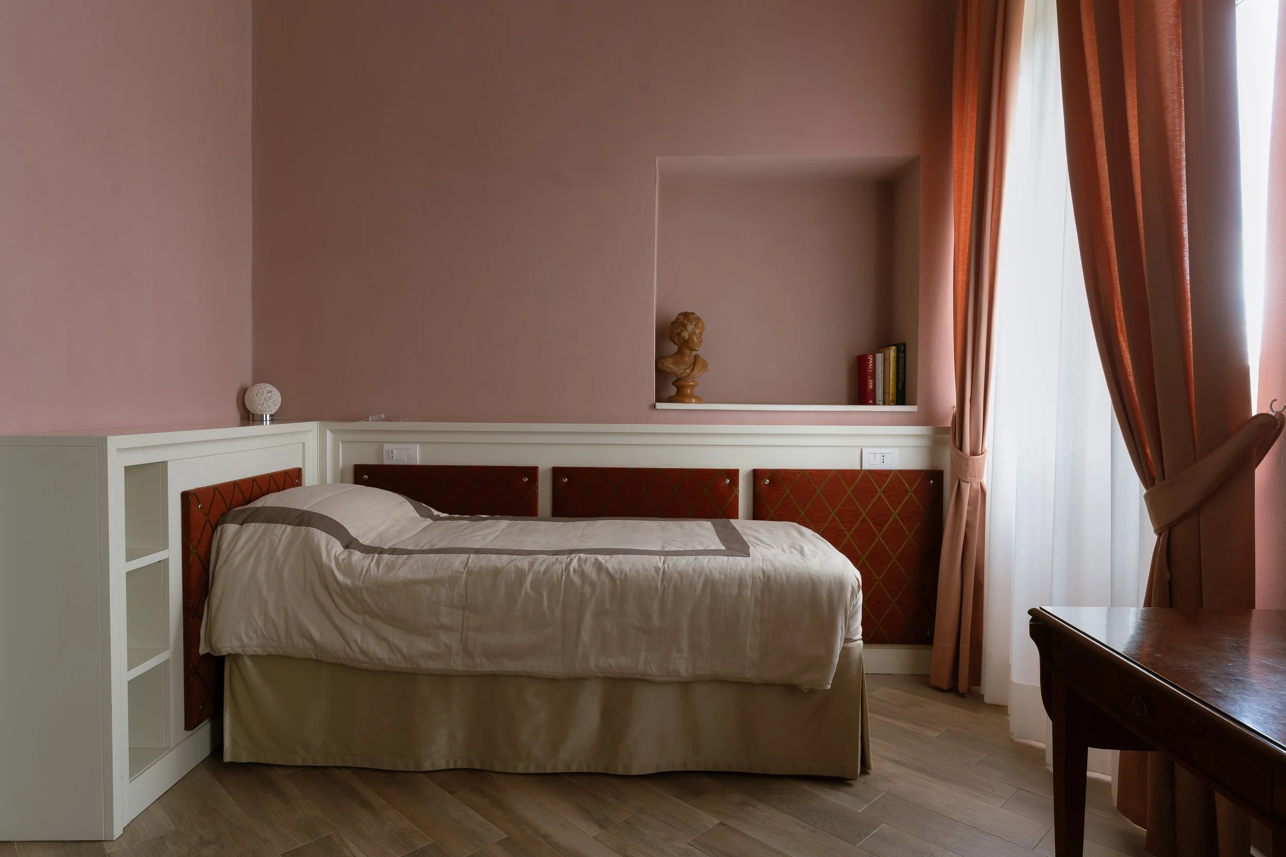 A bedroom with pink walls, a bed with a beige bedspread, and a built-in shelf with a bust sculpture and books. There is a large window with pink curtains to the right.