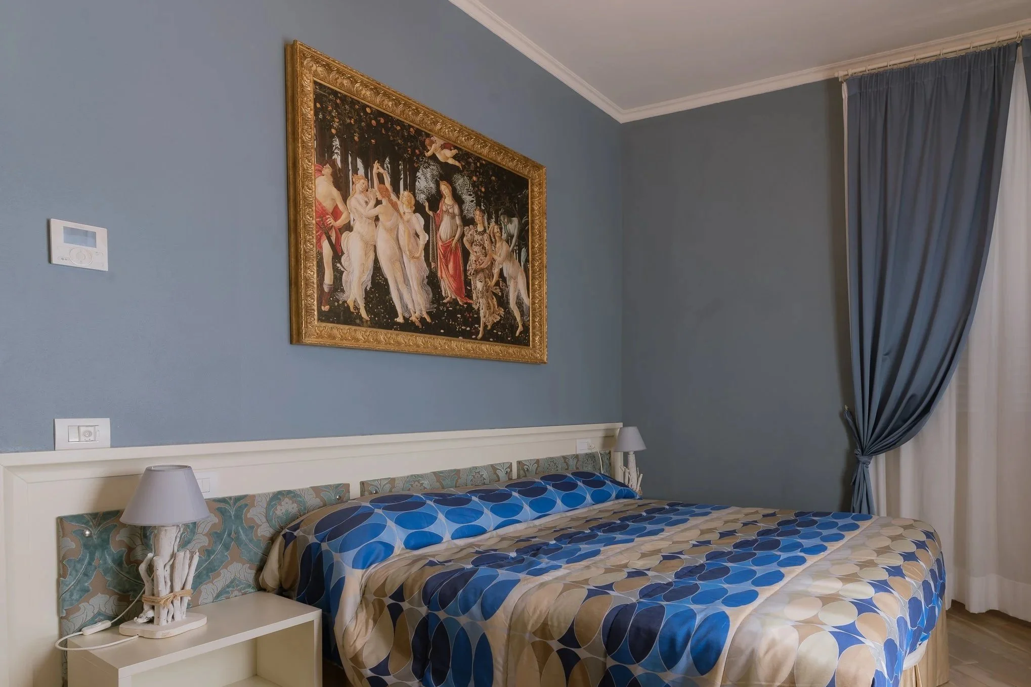 A bedroom with a bed that has a patterned bedspread in blue, beige, and cream colors. There are two lamps on bedside tables on either side of the bed. A large framed painting hangs on the blue wall above the bed. Blue curtains are drawn to the side, revealing a window with white sheer curtains.