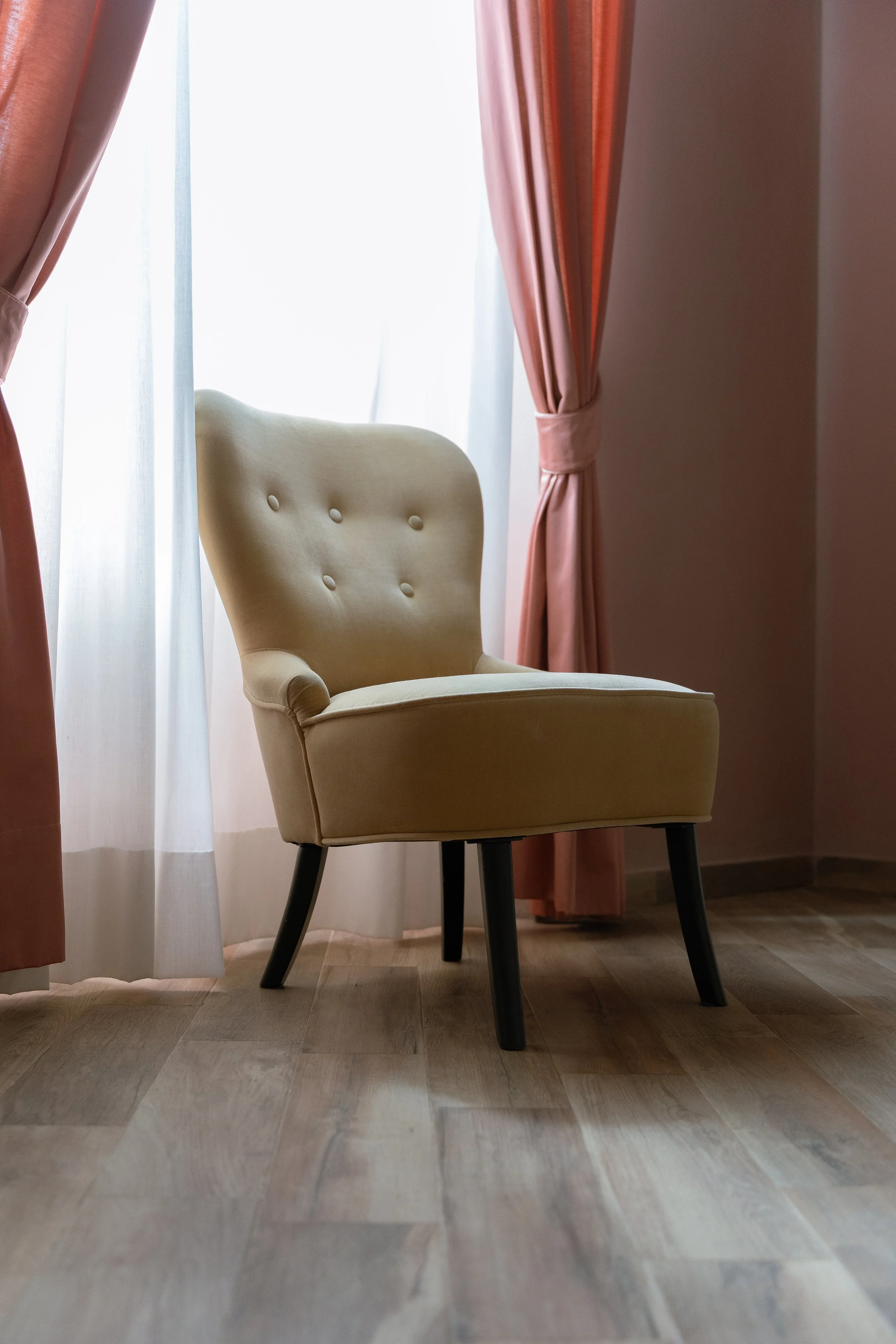 A beige upholstered armchair with black wooden legs, placed on a wooden floor near a window with white sheer curtains and pink drapes.