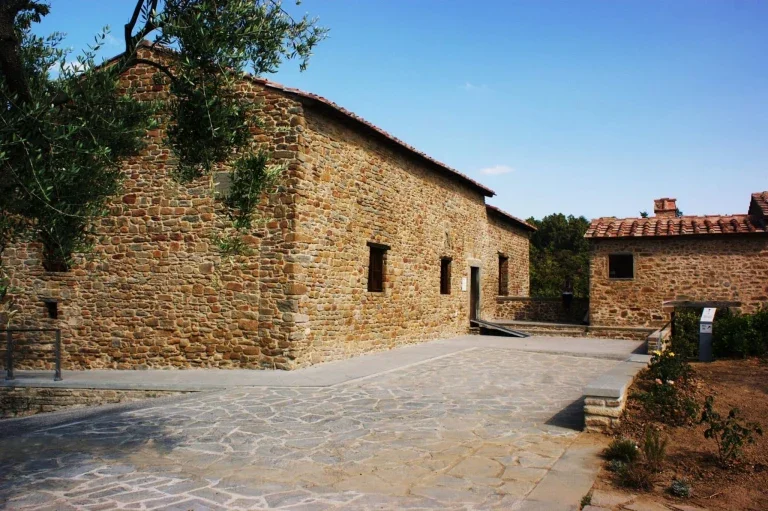 Stone buildings with small windows and tiled roofs in a rural setting, with a paved stone pathway and a tree in the foreground.