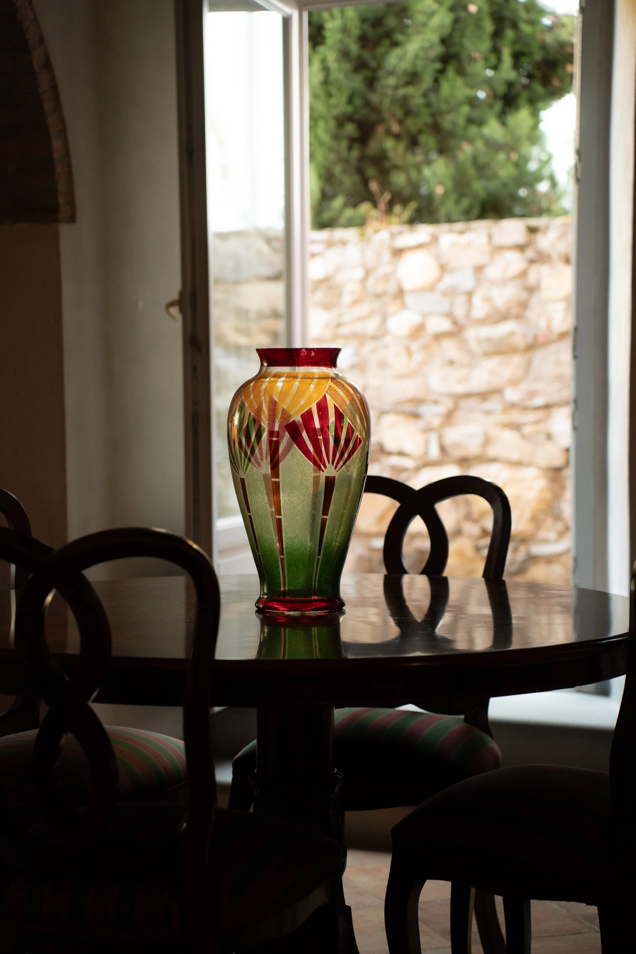 Colorful vase on a wooden dining table in a room with an open door showing a stone wall and green trees outside.