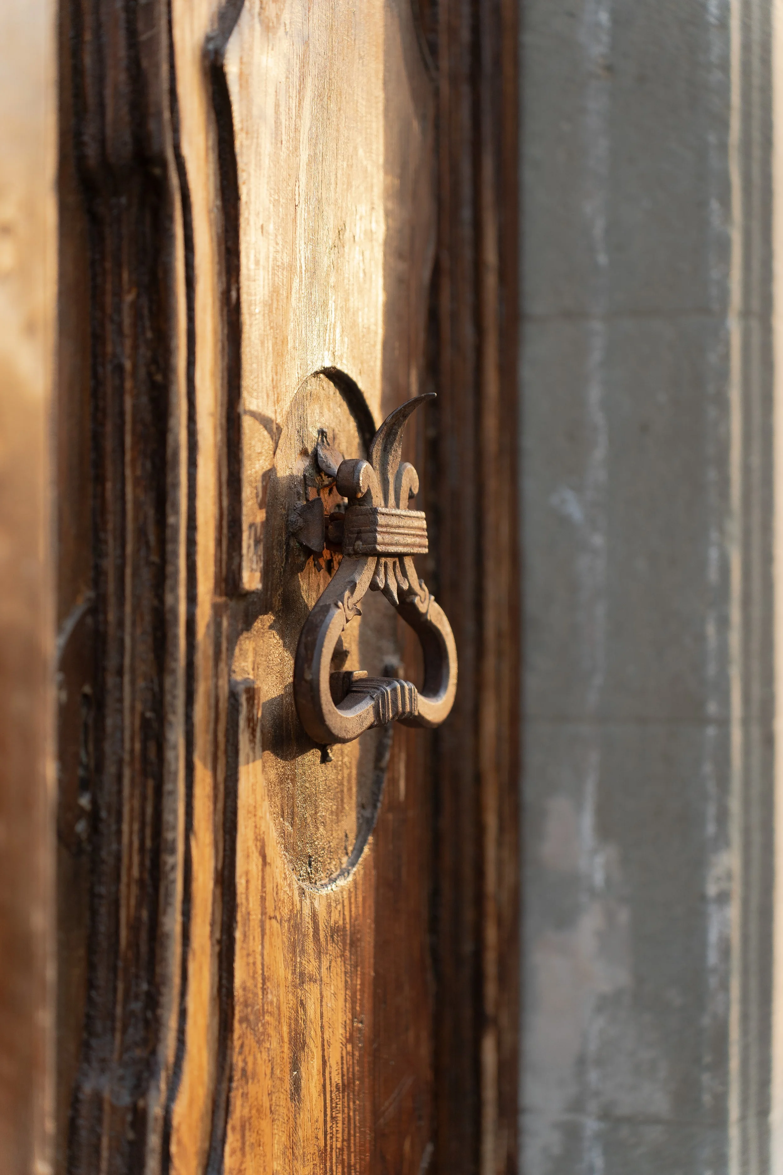 Close-up of a vintage iron door knocker mounted on a wooden door, with a cement wall visible to the right.