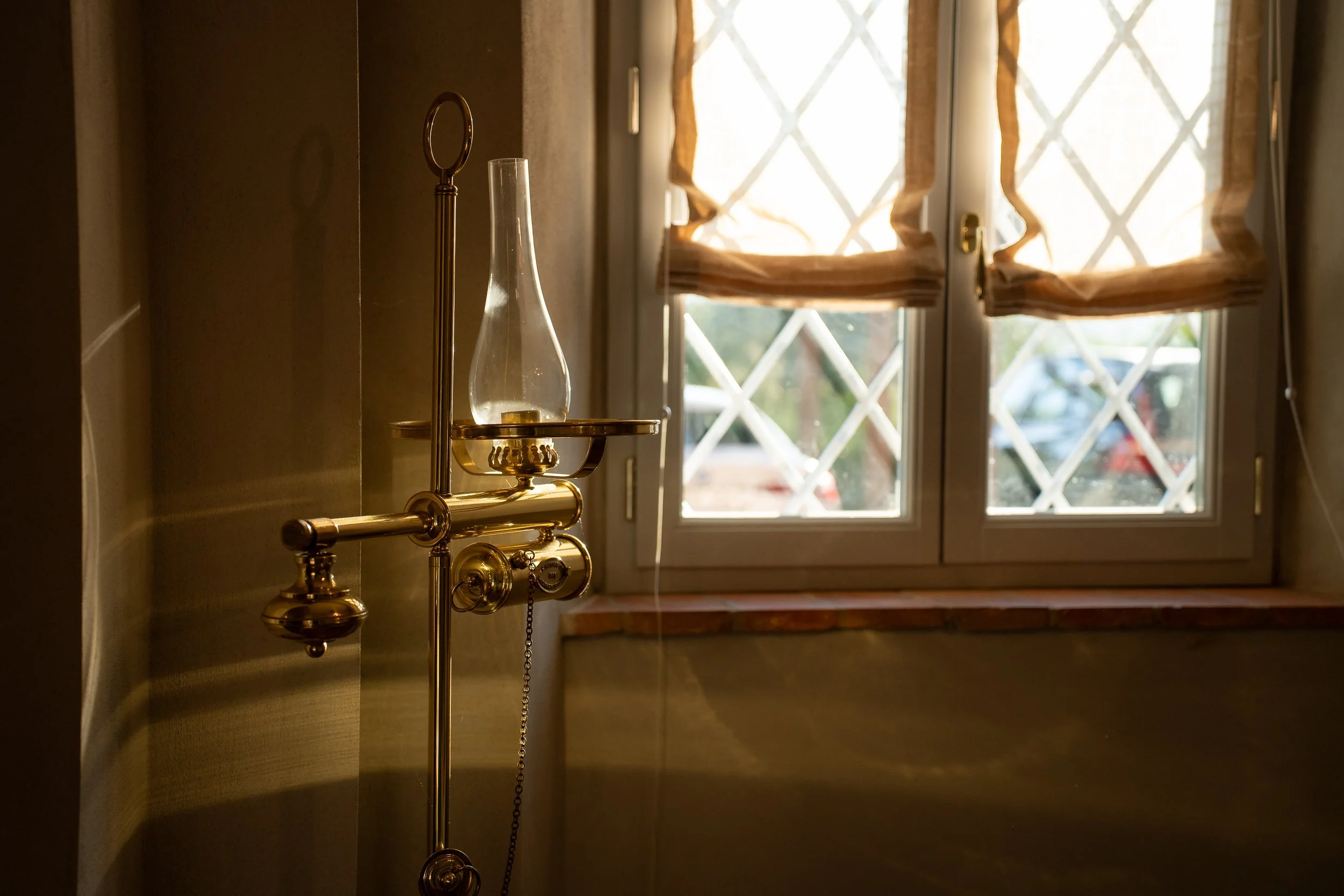 A vintage brass oil lamp with a glass chimney near a window with beige curtains, in a dimly lit room.