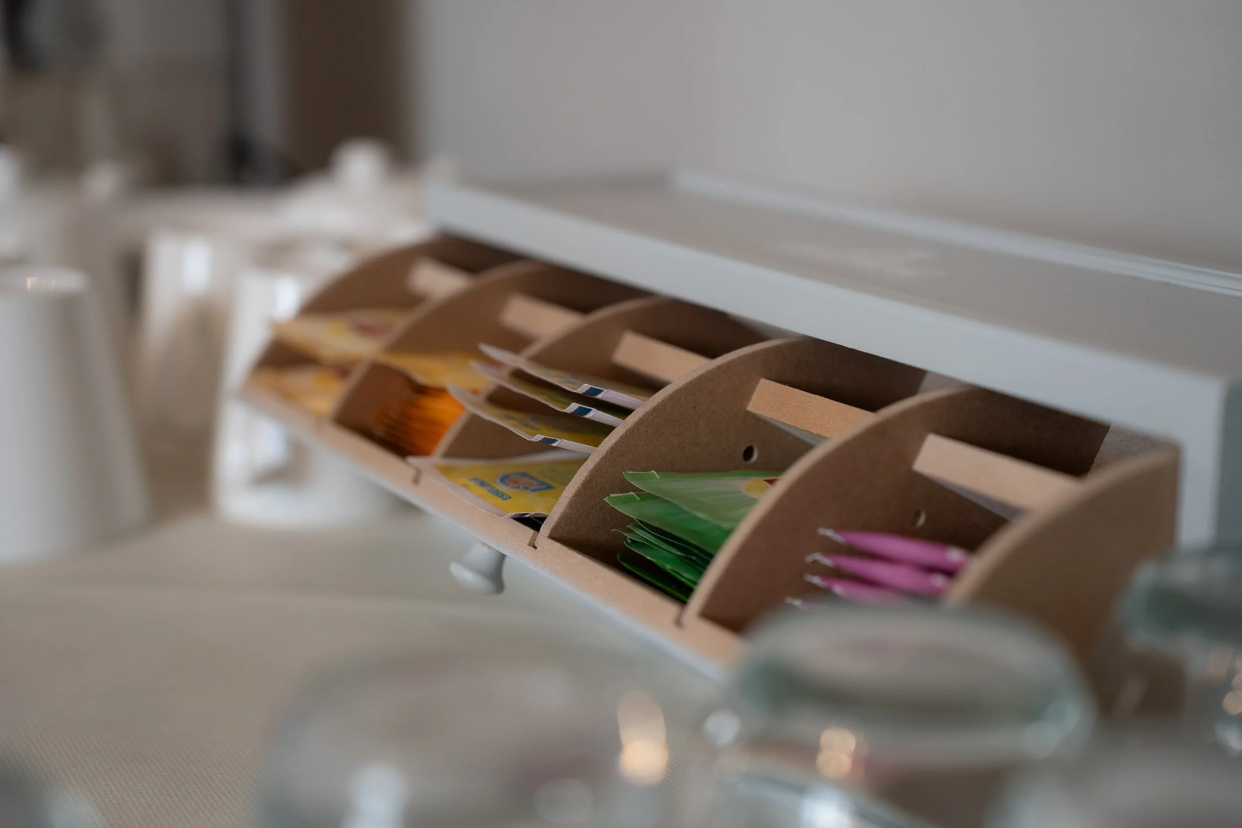 A wooden countertop with a spice and condiment rack filled with small packets of sugar, sweetener, and pink artificial sweetener. Blurred glassware is in the foreground.